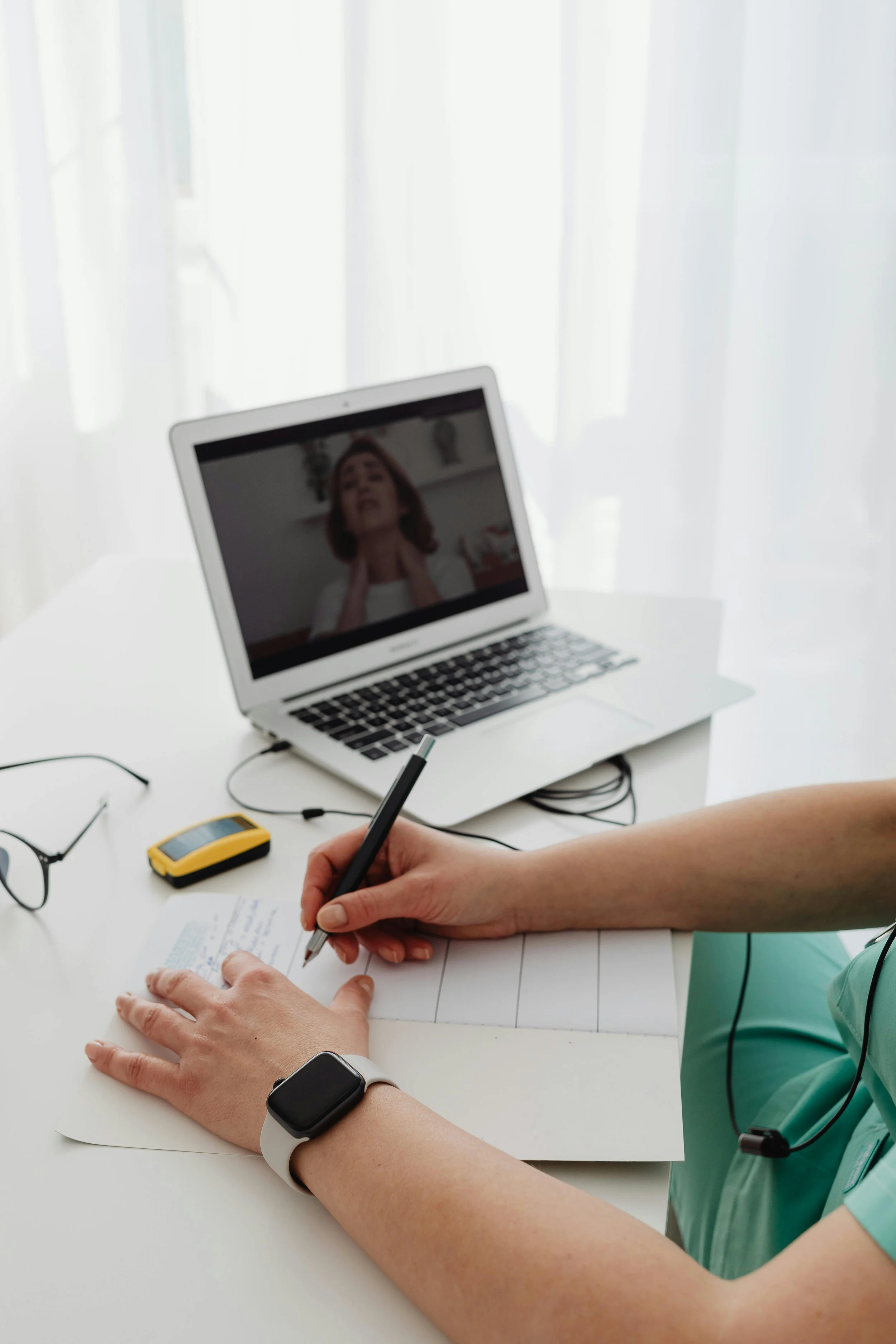 A person wearing a smartwatch writing notes on a paper at a desk, with a laptop displaying a video call in the background and a phone and glasses nearby.