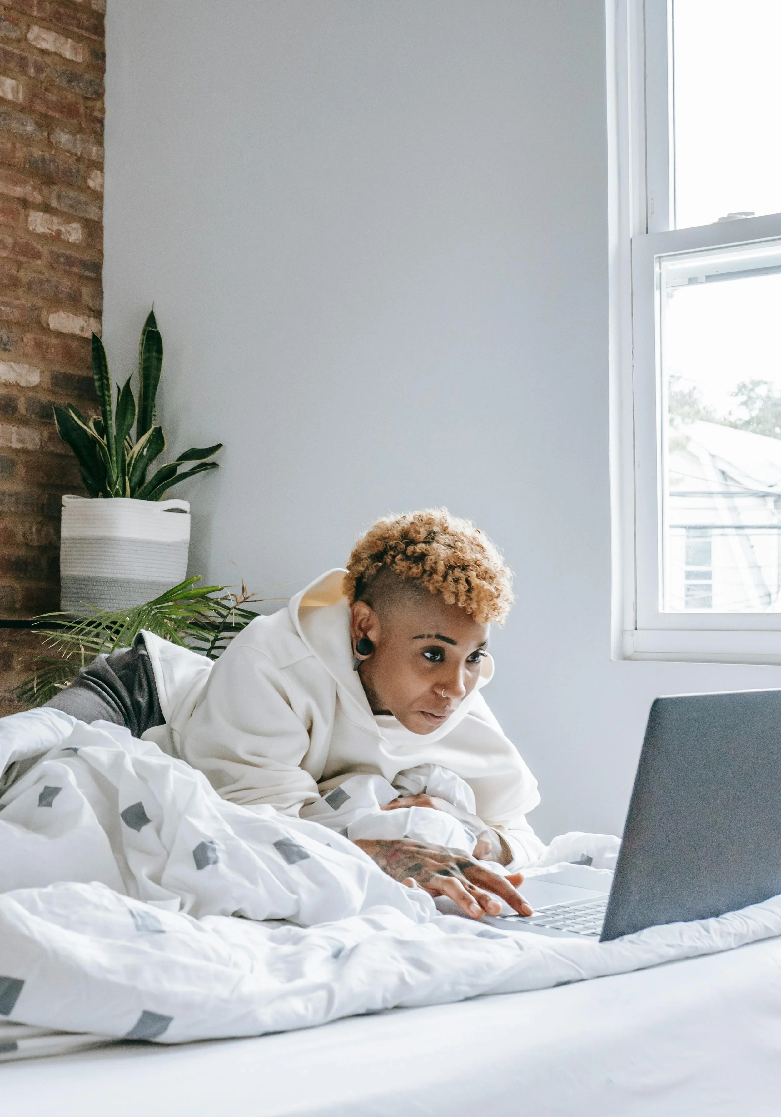 A person lying on their stomach on a bed, using a laptop while wrapped in a blanket, with a window and a potted plant in the background.