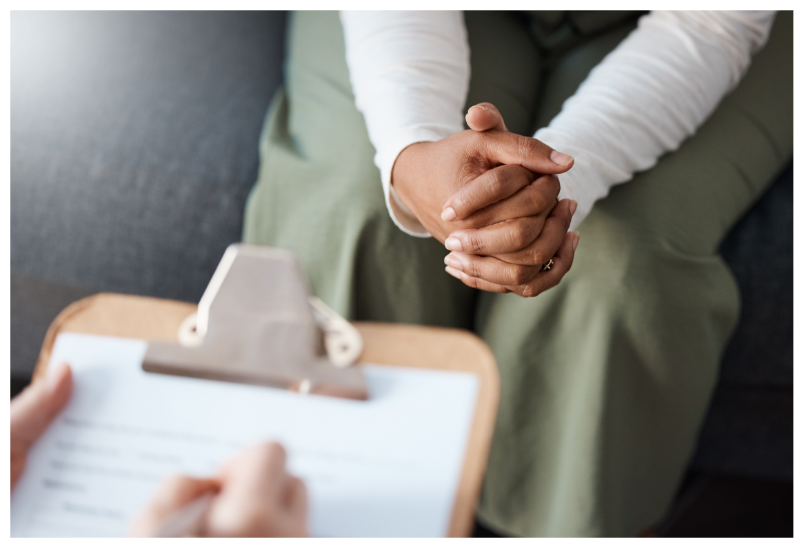 A person sitting on a couch with their hands clasped, talking to a professional or counselor who is holding a clipboard.