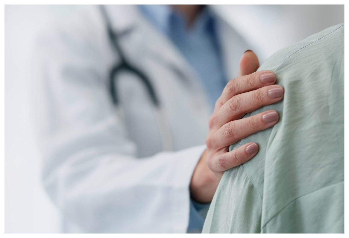 A close-up of a person in a white coat with a stethoscope placing a hand on another person's shoulder, suggesting a supportive or reassuring interaction.