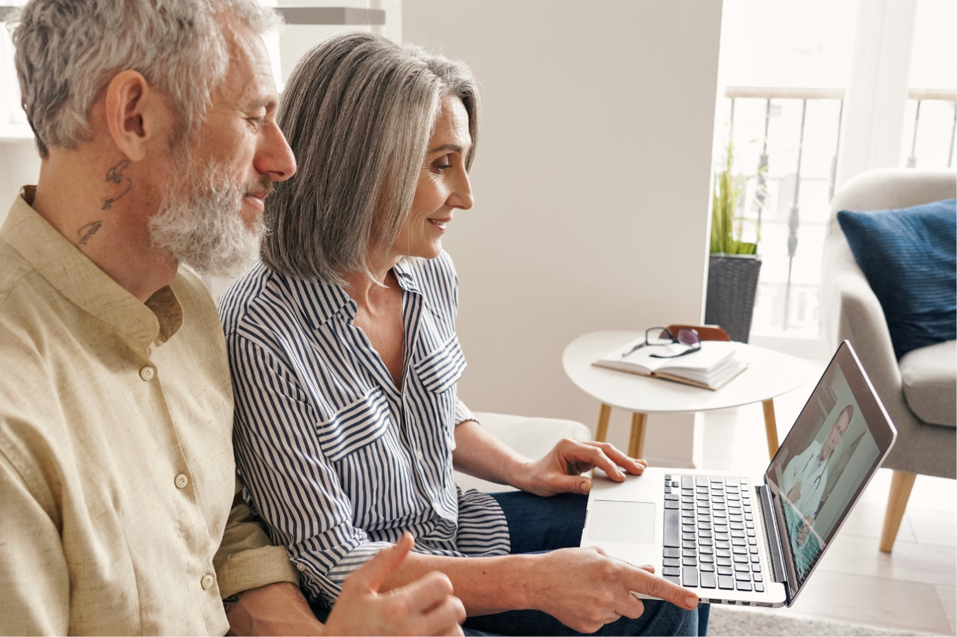 A man and a woman sitting side by side, smiling and looking at a laptop while the woman points at the screen, in a bright living room with a small table and books in the background.
