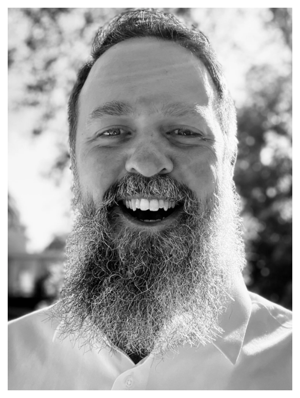 A smiling man with a full beard and mustache, wearing a collared shirt, outdoors with blurred trees in the background.