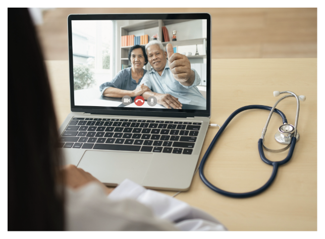 A laptop on a desk displaying a video call with an older man and a woman smiling at the camera, while a person sits nearby and a stethoscope rests on the desk next to the laptop.