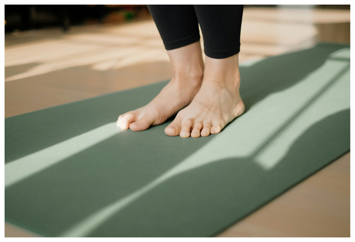 A close-up of a person's bare feet standing on a yoga mat, with soft lighting casting shadows across the mat.