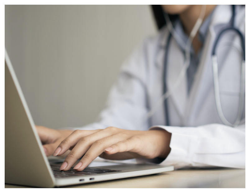 A close-up of a person in a white coat with a stethoscope around their neck, typing on a laptop while seated at a desk.