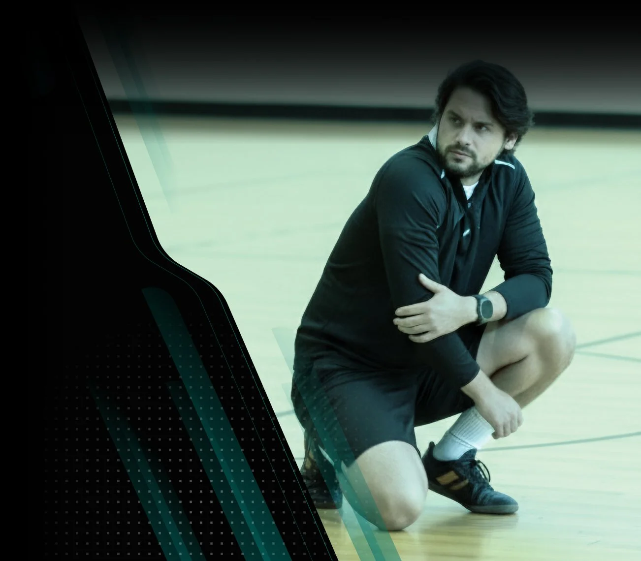 A man with dark hair and stubble, sitting on a gymnasium floor, wearing athletic clothing and a watch, appearing to stretch or rest after exercising.