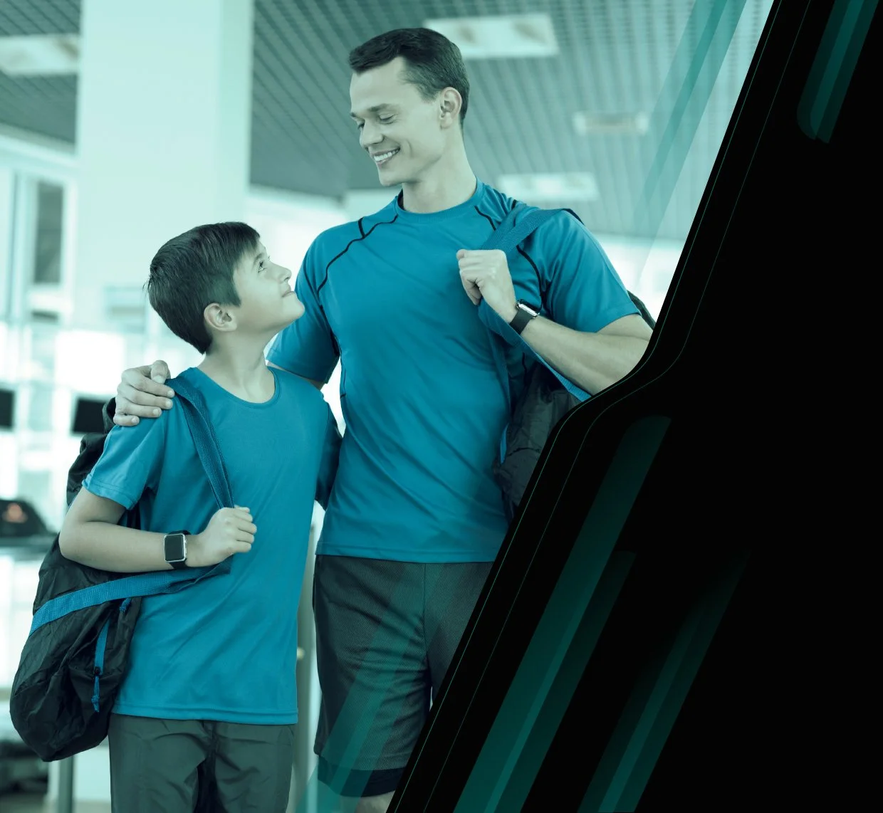 A man and a young boy, both dressed in athletic clothing and carrying backpacks, are smiling at each other at an airport or similar public transport location.