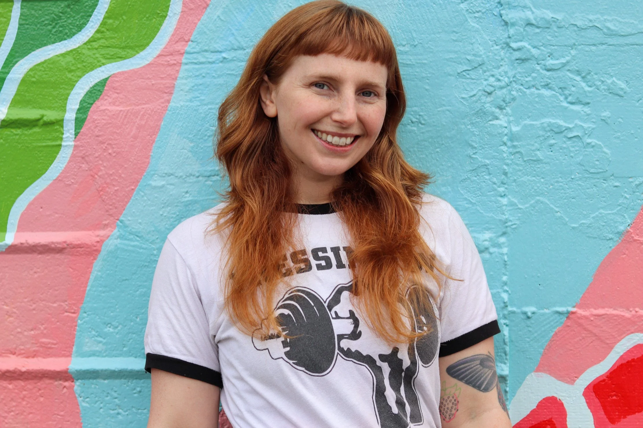 A woman with long red hair smiling in front of a colorful painted wall.