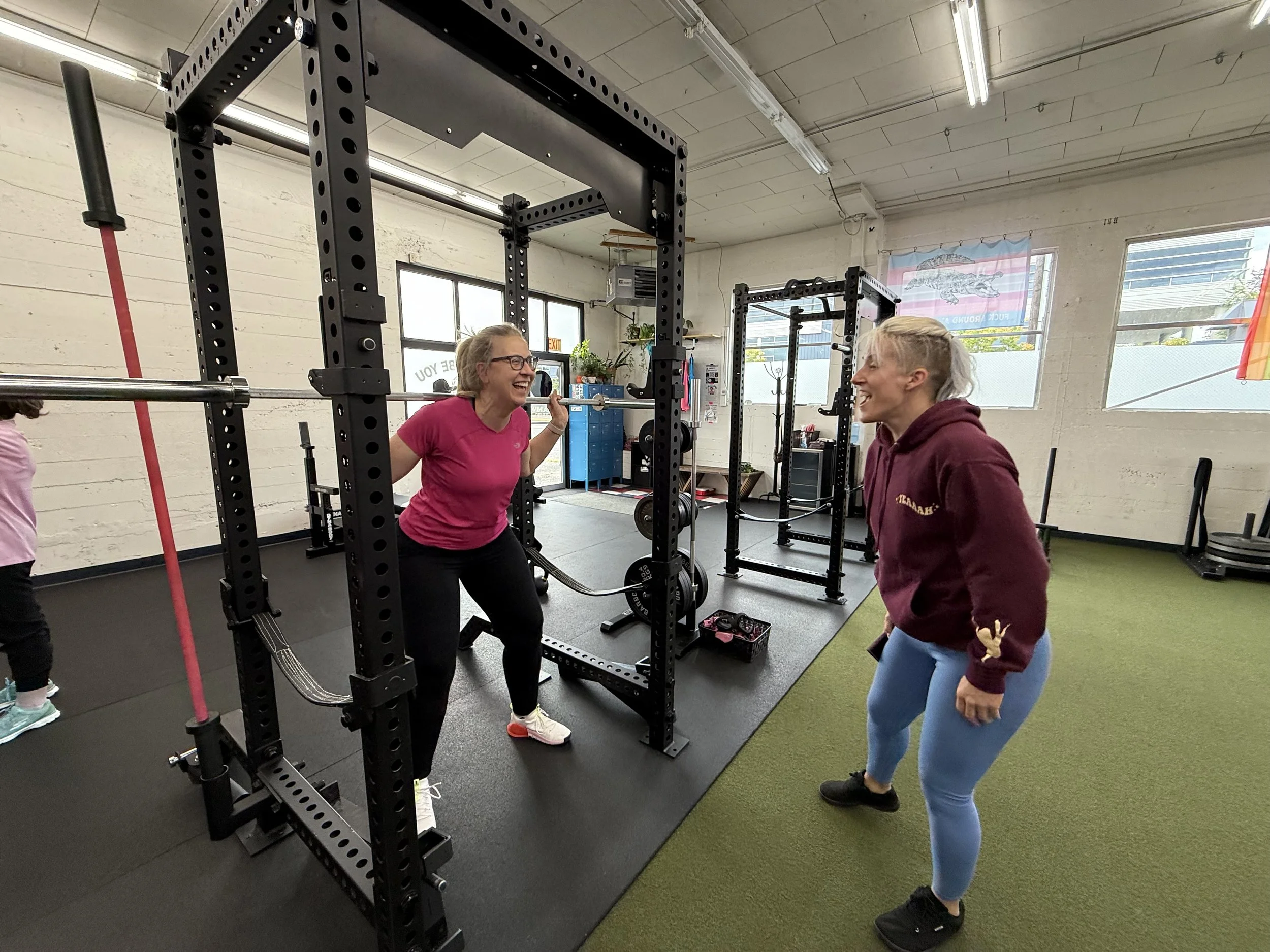 Two women smiling and talking in a gym, with one doing squats under a power rack and the other standing nearby.