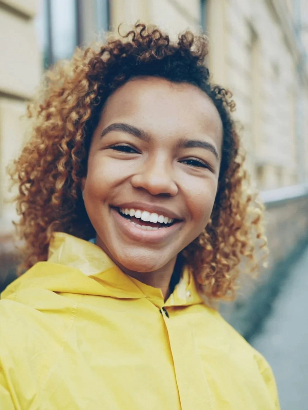Close-up of a woman smiling, wearing a yellow raincoat, with curly hair, outdoors near a building with large windows.