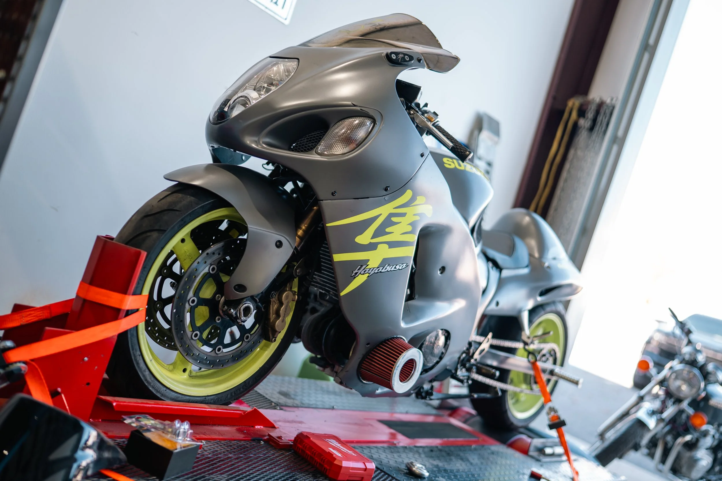 A sleek, matte gray sport motorcycle with yellow accents and decals, mounted on a red stand indoors, with tools and another motorcycle visible in the background.