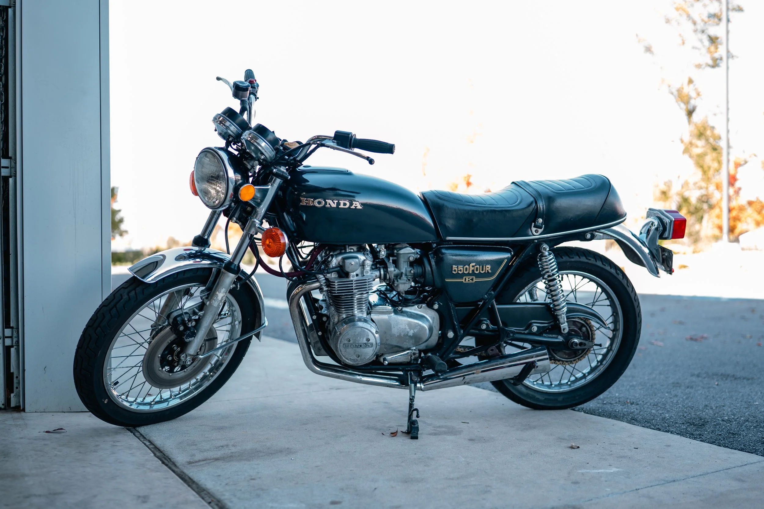 A vintage black Honda 550 Four motorcycle parked on a concrete surface with a background of trees and sky.