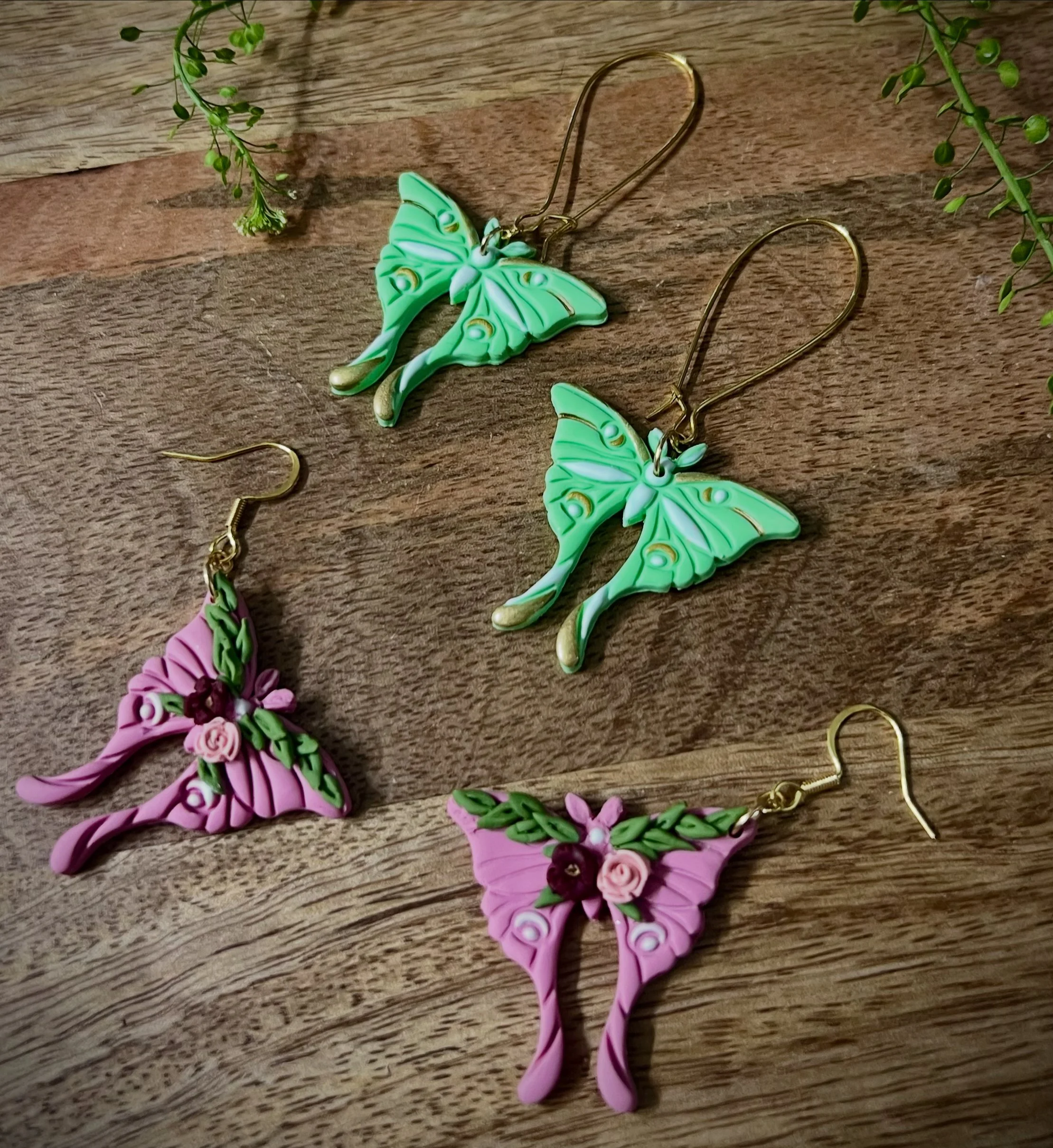 Colorful butterfly/moth-shaped earrings, two green ones and two pink ones, arranged on a wooden surface with some greenery around.