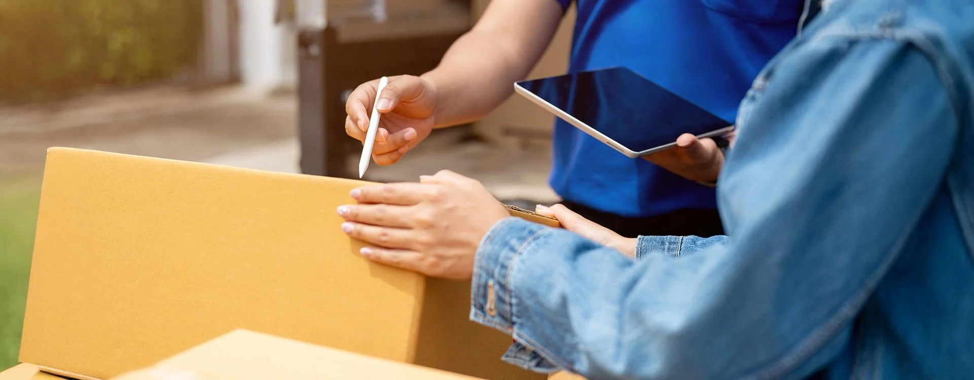 Two people are handling a yellow cardboard package; one person is holding a tablet, and the other is holding a stylus.