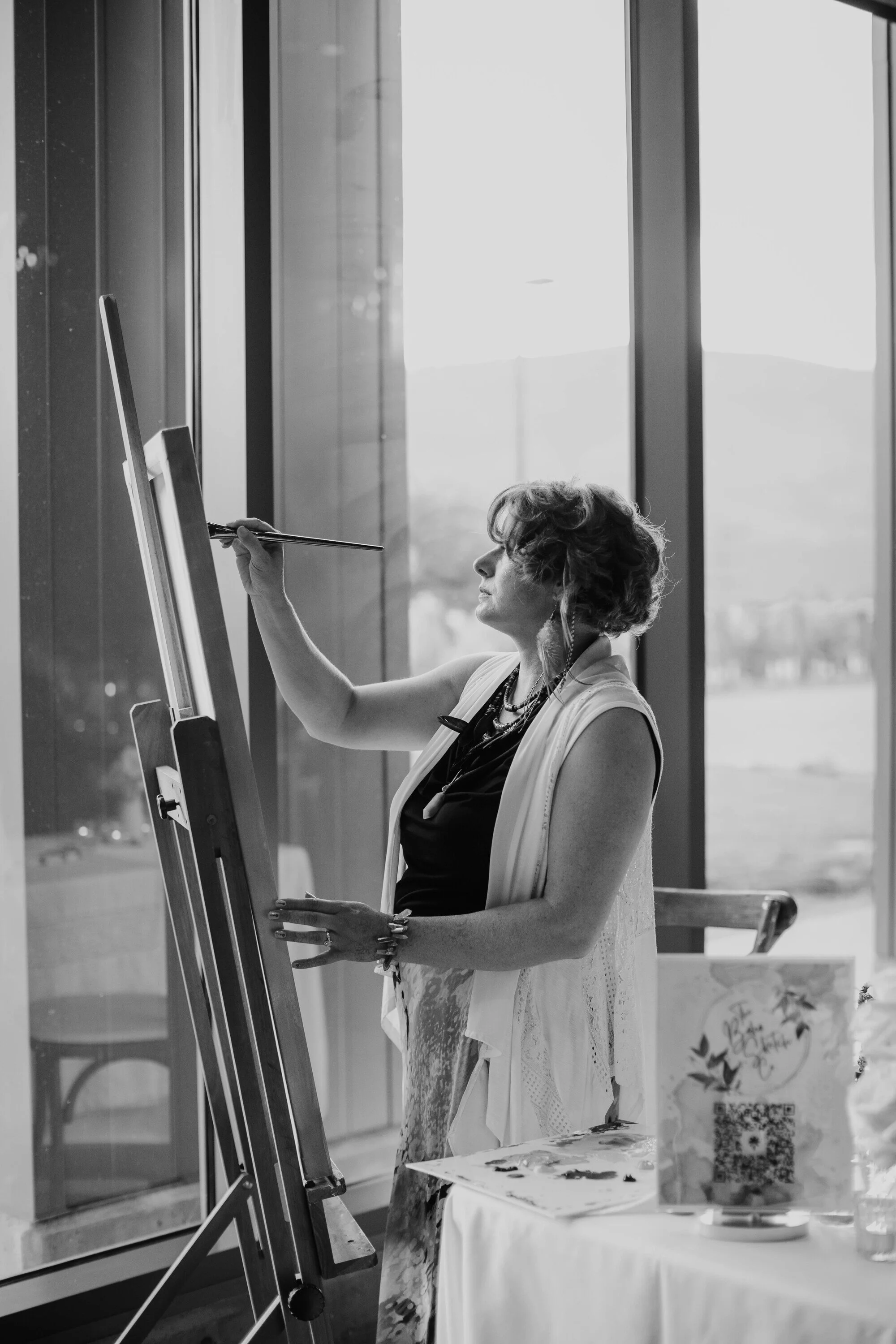 A woman standing at an easel, drawing, inside a room with large windows showing an outdoor landscape.