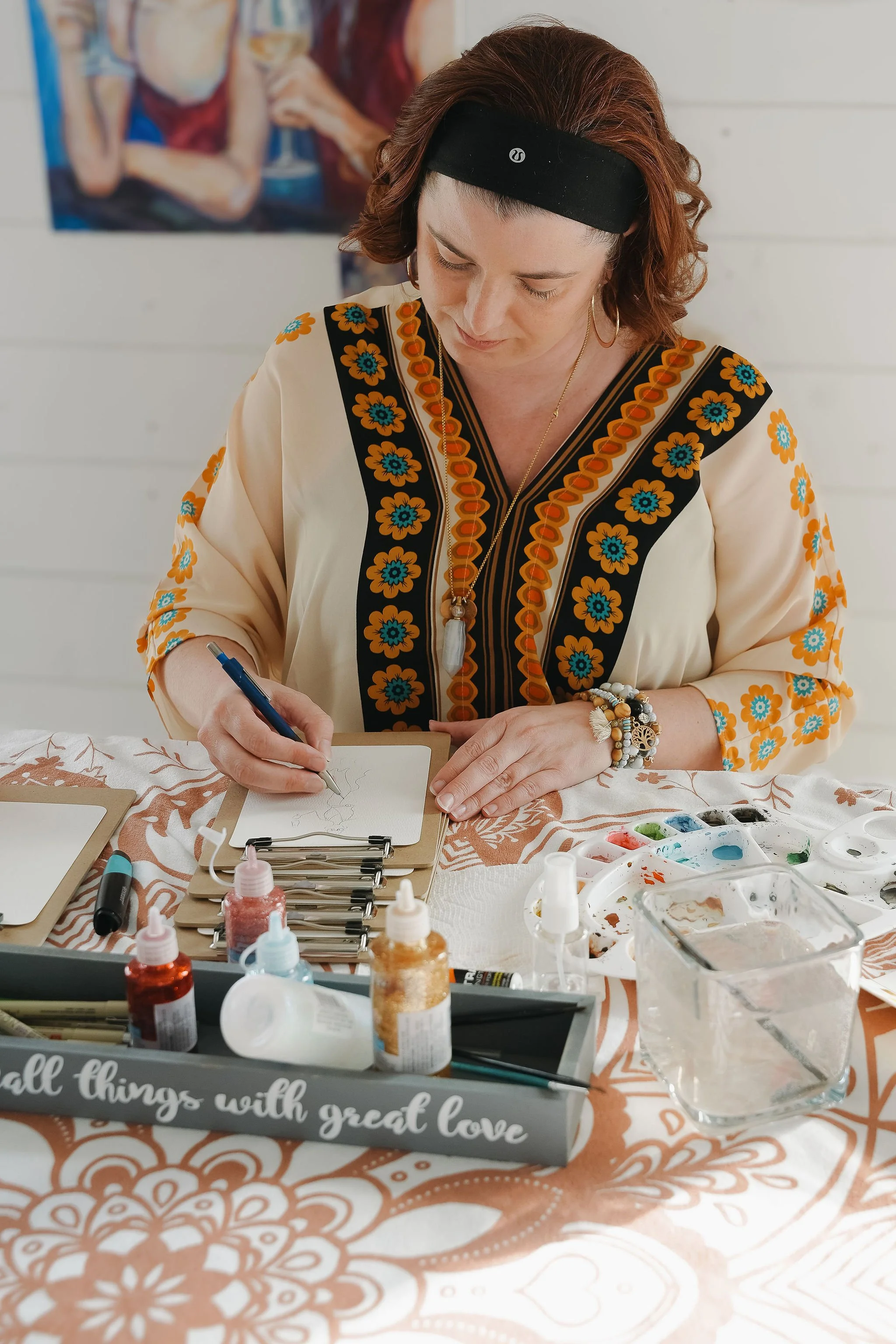 Shannon, the artist at Boho Sketch Co. is sketching on paper, surrounded by art supplies like paint, brushes, and water, with a decorative tray on the table that reads 'all things with good love.'