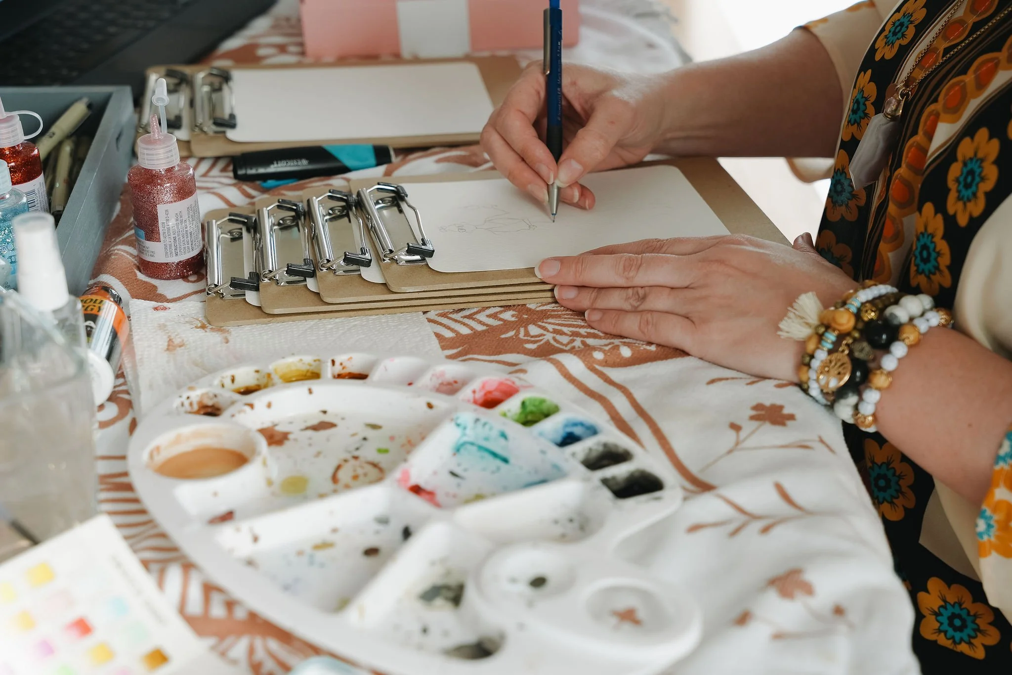 Shannon, the artist at Boho Sketch Co. drawing on a clipboard, surrounded by watercolour paints, brushes, and crafting supplies on a table.