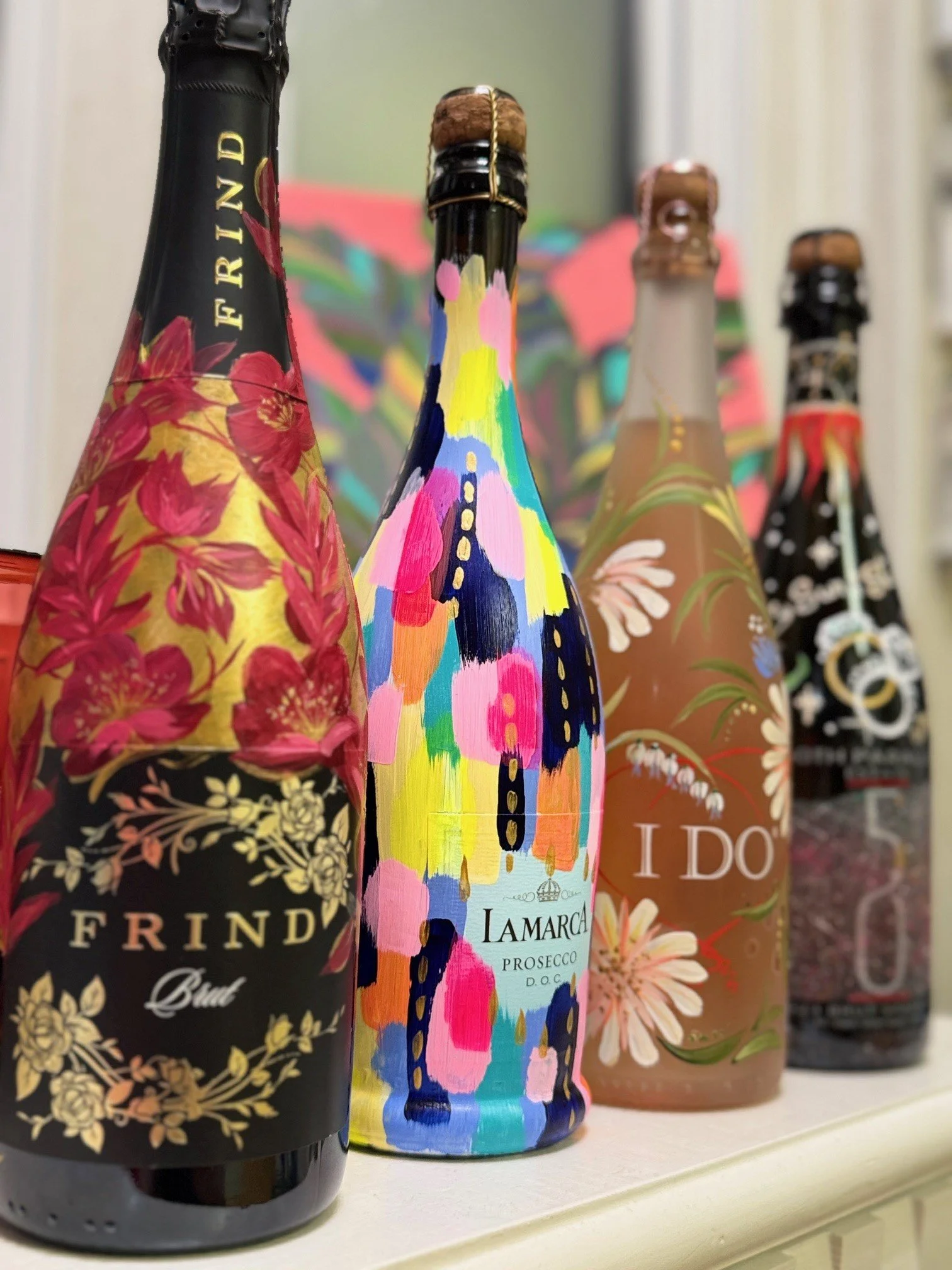 Four custom painted wine bottles with colourful, artistic labels on a shelf.