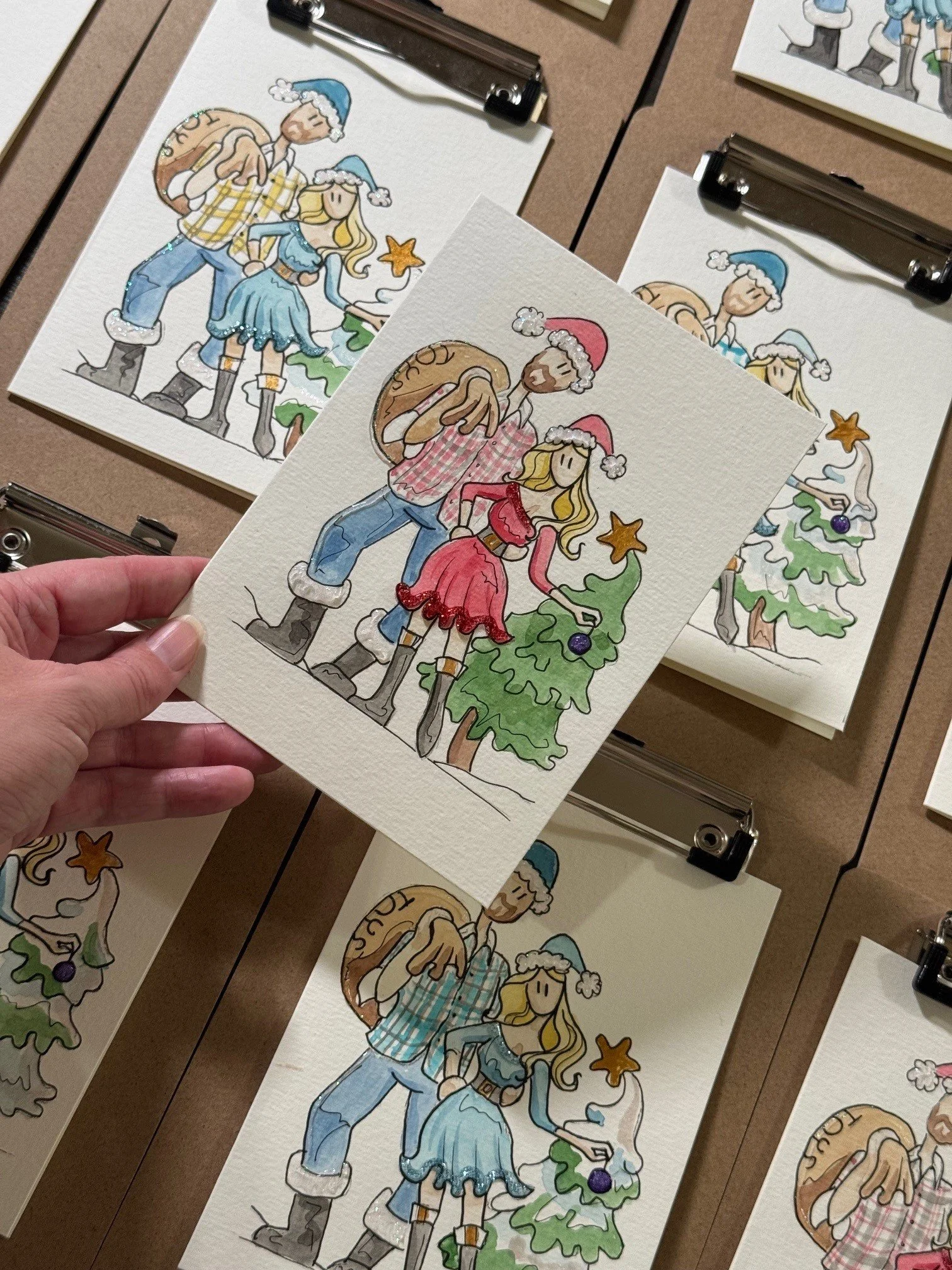 Watercolor illustrations of a couple dressed in Christmas-themed clothing, standing next to a decorated Christmas tree. The man is wearing a Santa hat, checkered shirt, jeans, and boots, carrying a large sack. The woman is in a red dress, Santa hat, and boots, with long blonde hair.