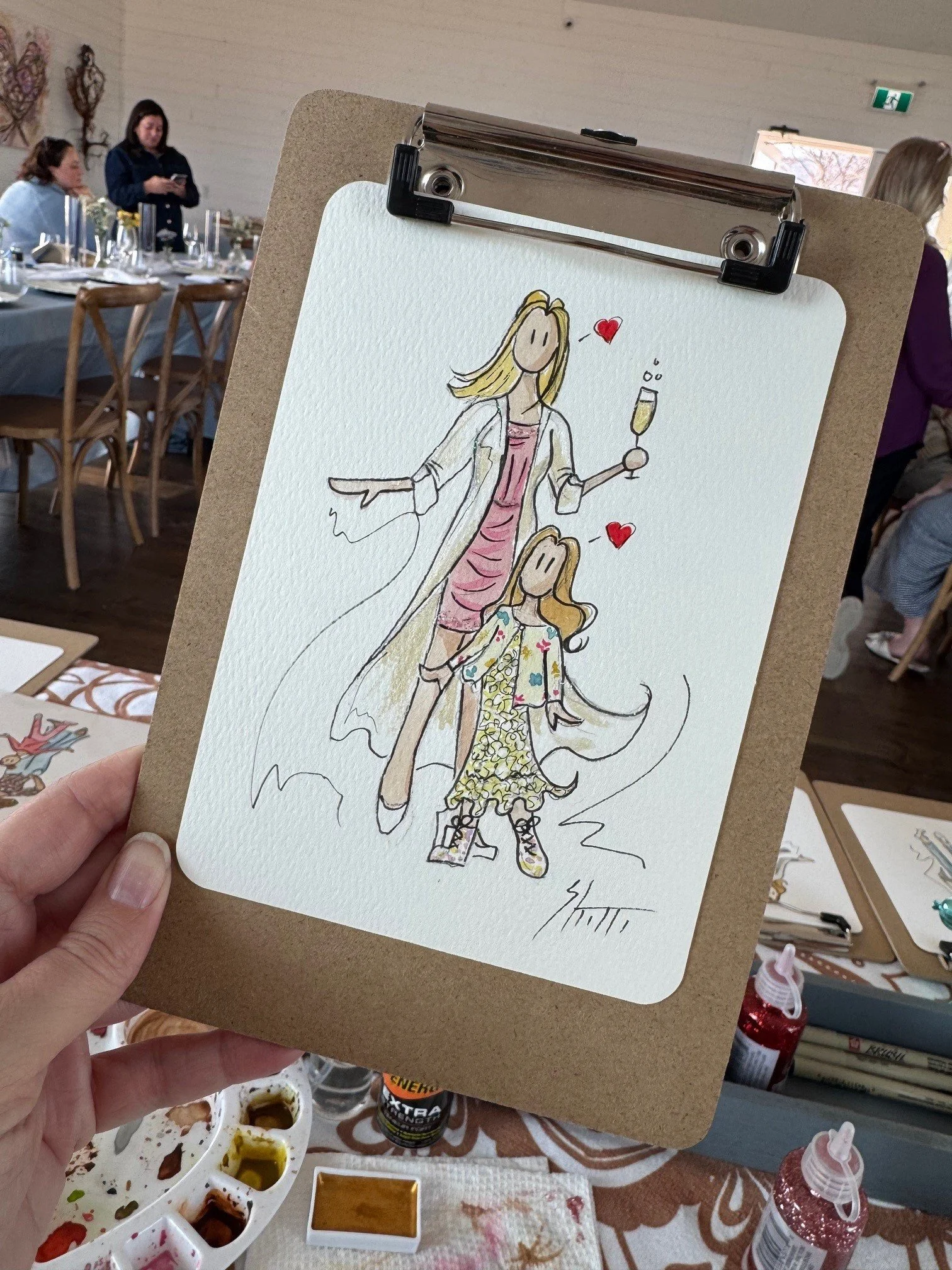 A hand holding a sketch of two women, one older and one younger, with hearts near their heads. The older woman has blonde hair and is holding a champagne glass, while the younger woman has long, visible hair and is wearing a floral dress. The sketch is on a piece of paper attached to a clipboard, with a background showing a room with people, tables, and art supplies.