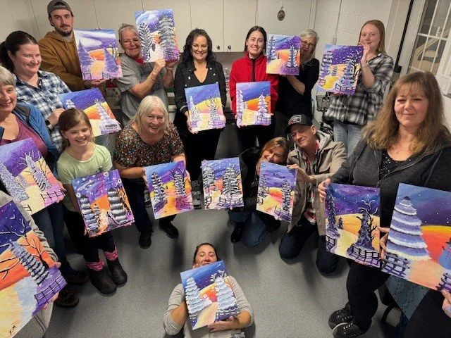 Group of people holding up colorful winter landscape paintings with snow-covered trees.
