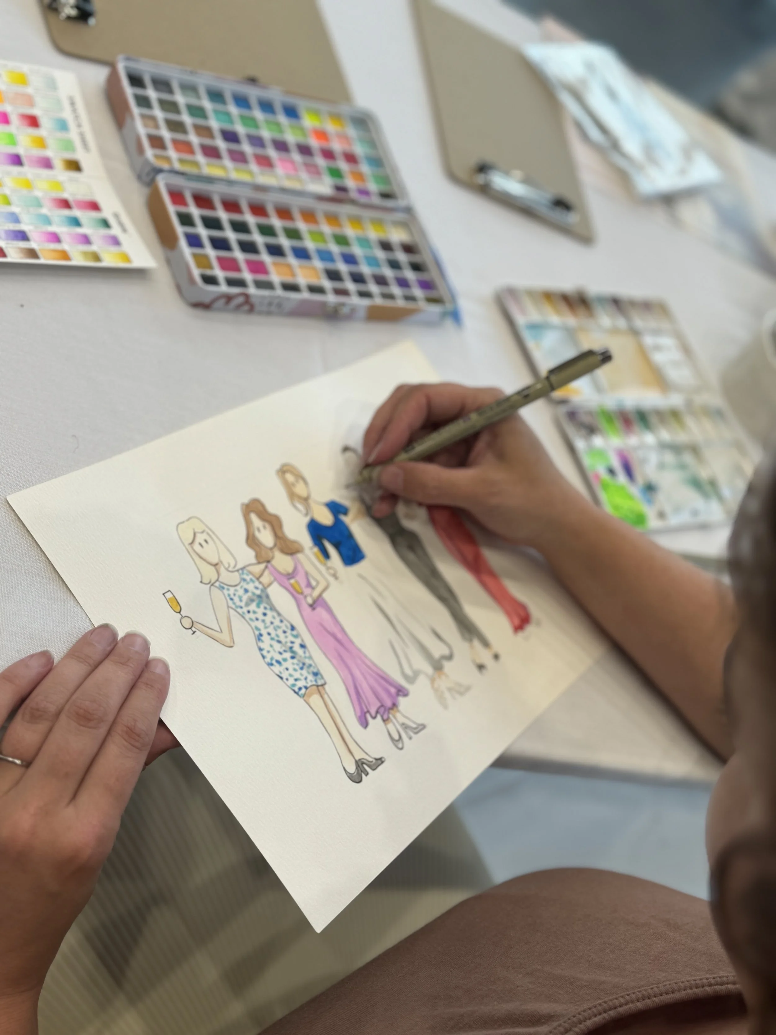 A person is creating a watercolor illustration of four women dressed for a celebration, with makeup, accessories, and dresses, at a table with watercolor paints and brushes.