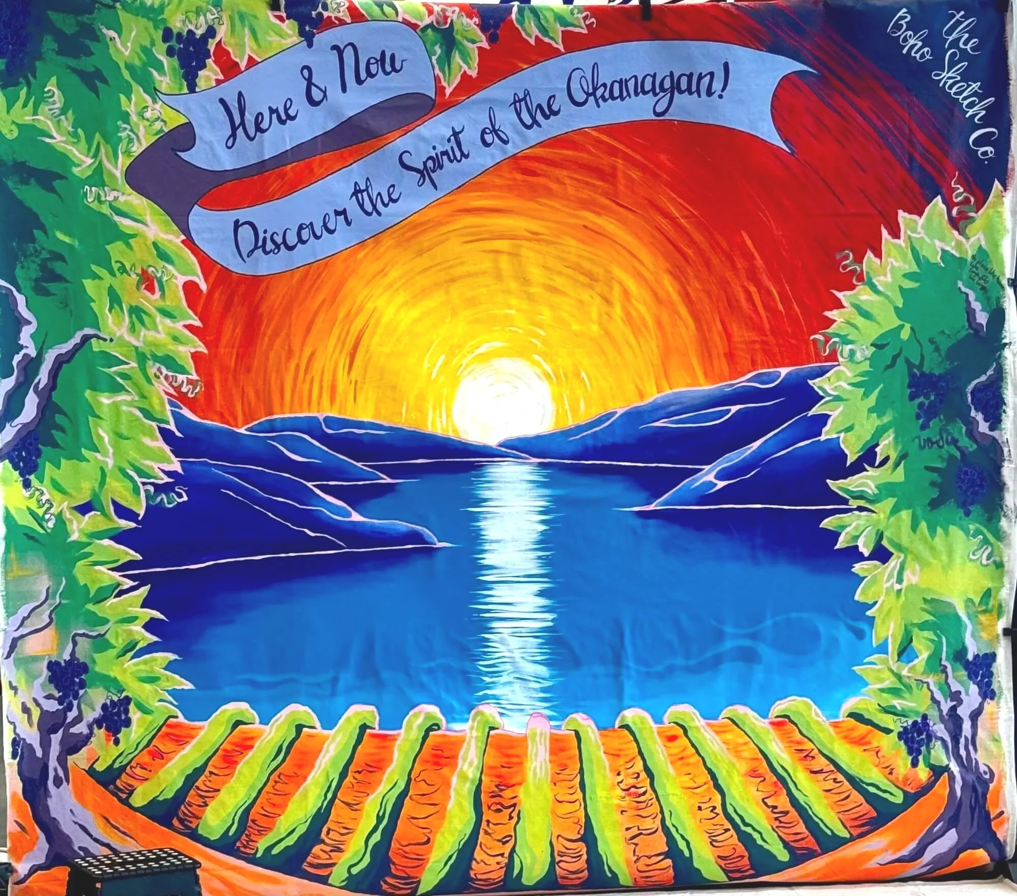 A vibrant painting of a sunset over a river, surrounded by mountains and framed with grapevines. A blue banner reads, 'Here & Now Discover the Spirit of the Okanagan!' in cursive. The scene is colorful with bright orange, yellow, green, and blue hues