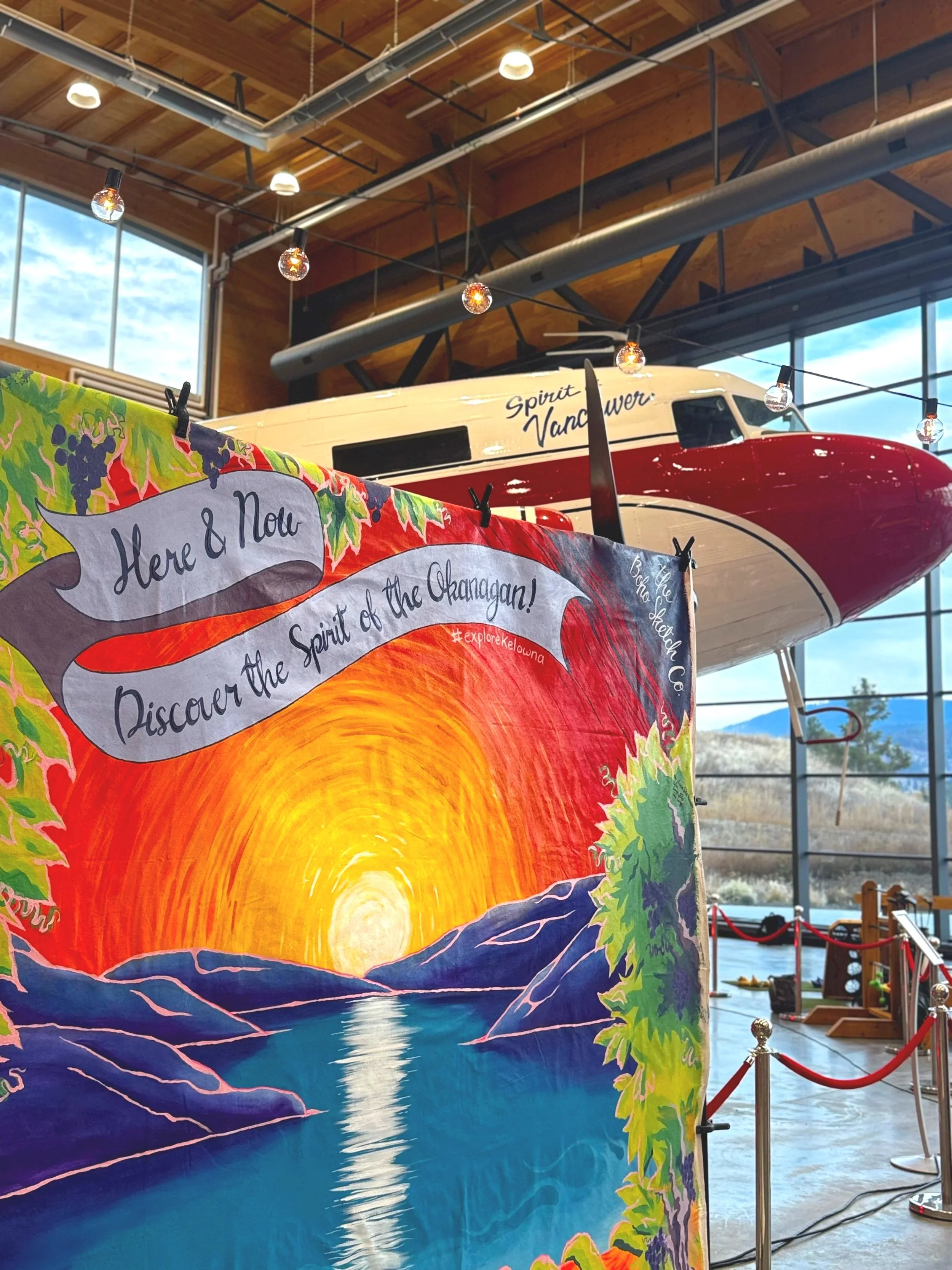 Interior of a museum with a large red and white airplane mounted on the wall and a colorful artwork featuring a sunset over mountains and water in the foreground.