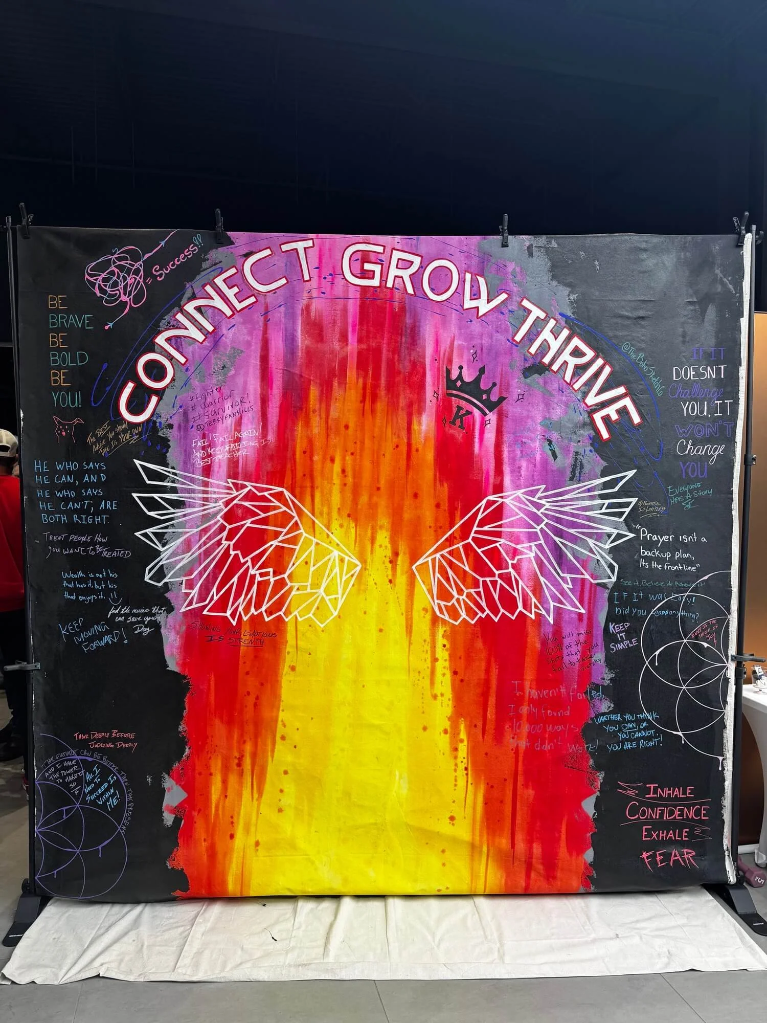 Colorful backdrop with the words "Connect, Grow, Thrive" at the top, a crown with the letter "K" in the center, and white outlined angel wings; the backdrop features handwritten notes and motivational quotes on a black background.