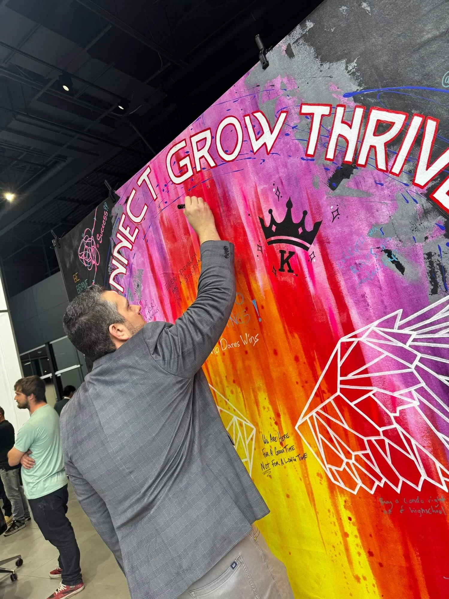 A man in a gray blazer writes on a colorful mural with the words 'Connect Grow Thrive' in large pink letters. The mural features a purple crown and a white geometric butterfly design.