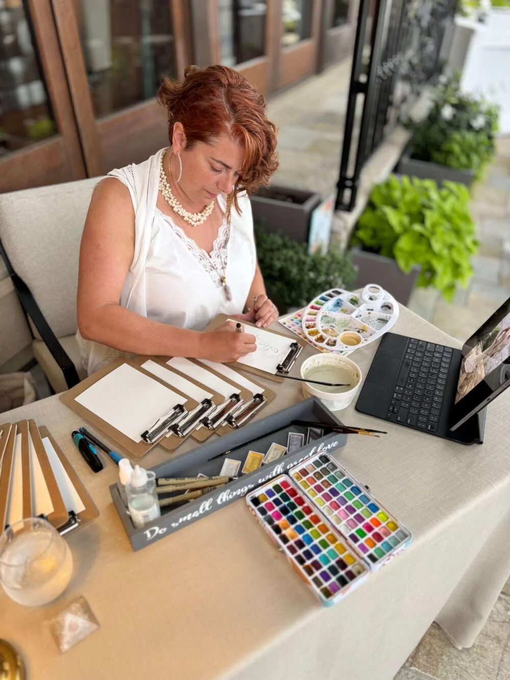 The artist at Boho Sketch Co. is sitting at a table painting on a clipboard with watercolor supplies, including a palette, brushes, and watercolors, with a laptop and plants in the background.