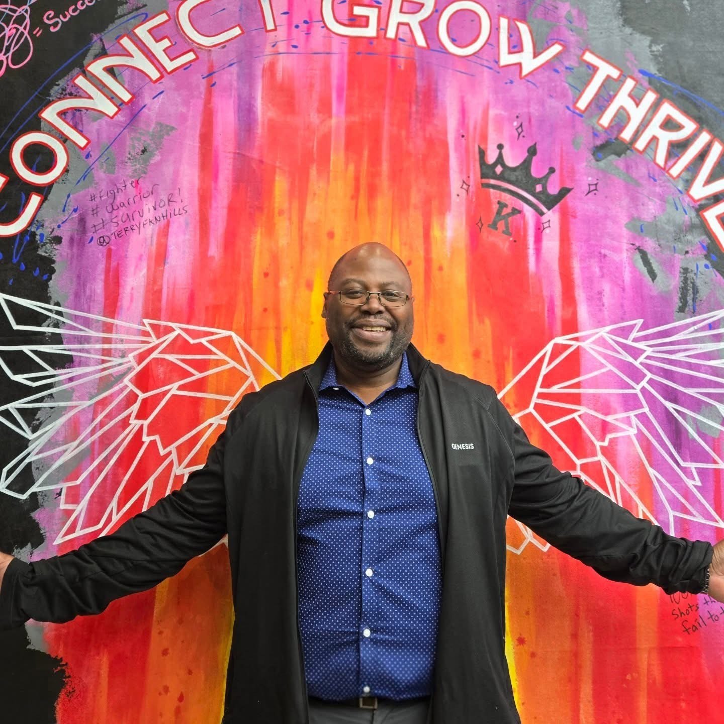 A smiling man standing in front of a colorful mural with angel wings and the words 'Connect, Grow, Thrive'.