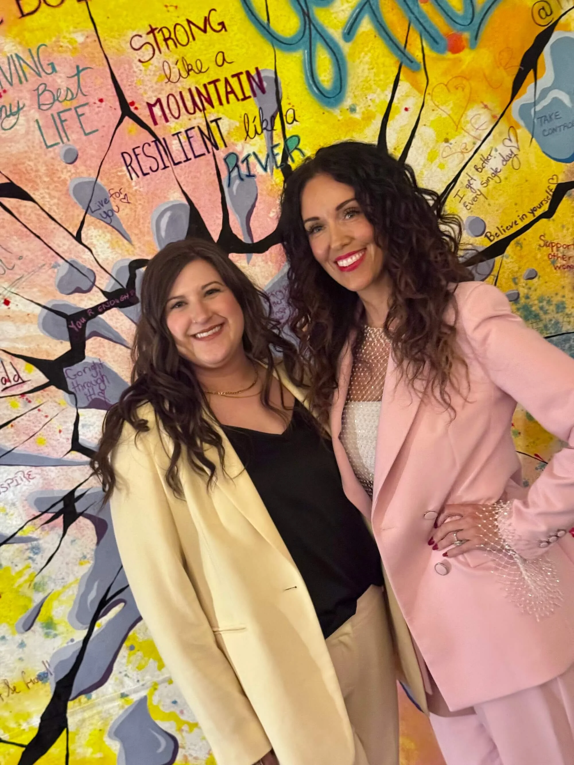 Two women smiling in front of a colorful mural painted live at a corporate event.