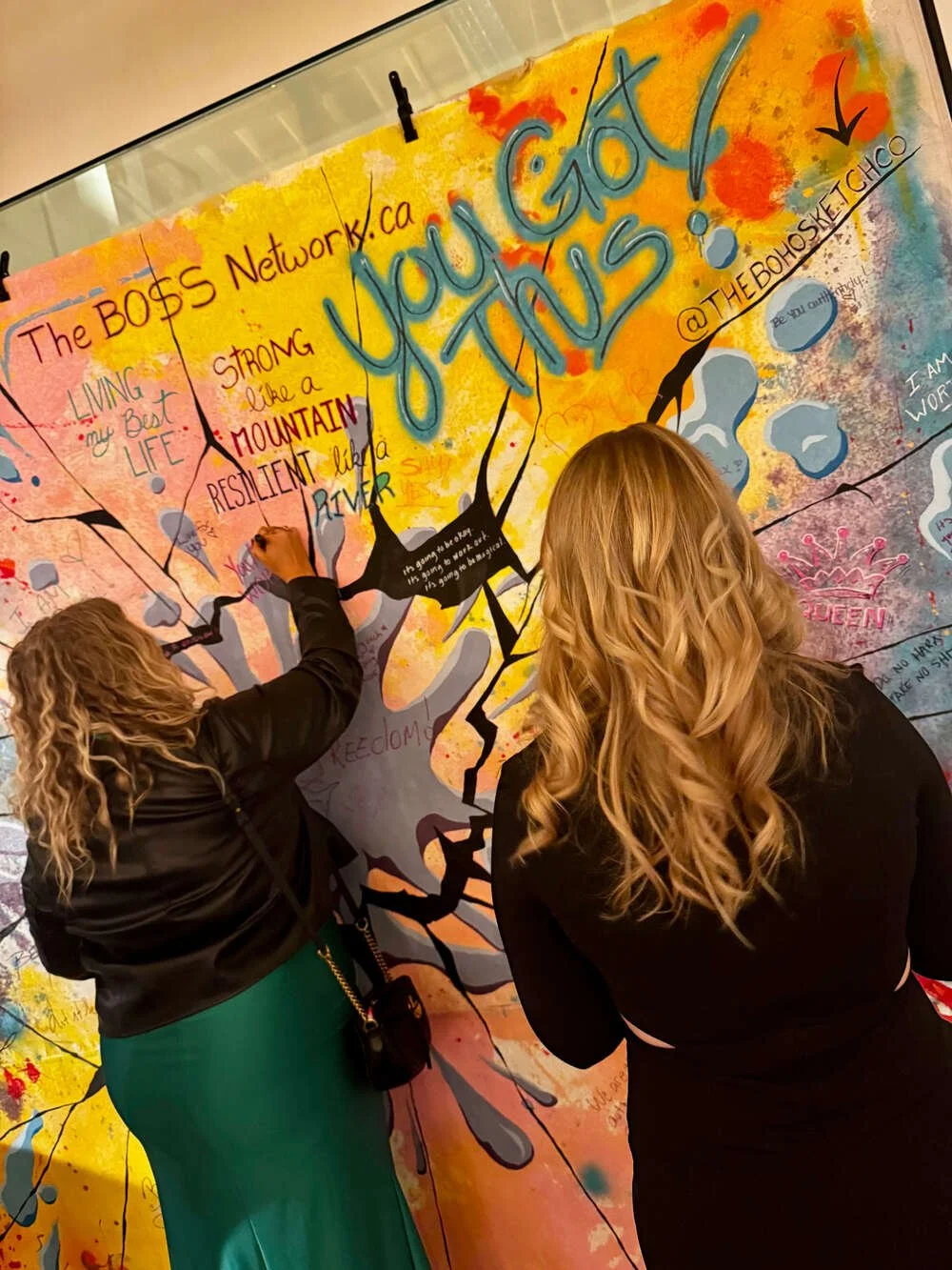 Two women writing on a colorful, artistic mural with handwritten messages and drawings, which includes the phrase 'What you think!' in large, blue letters.