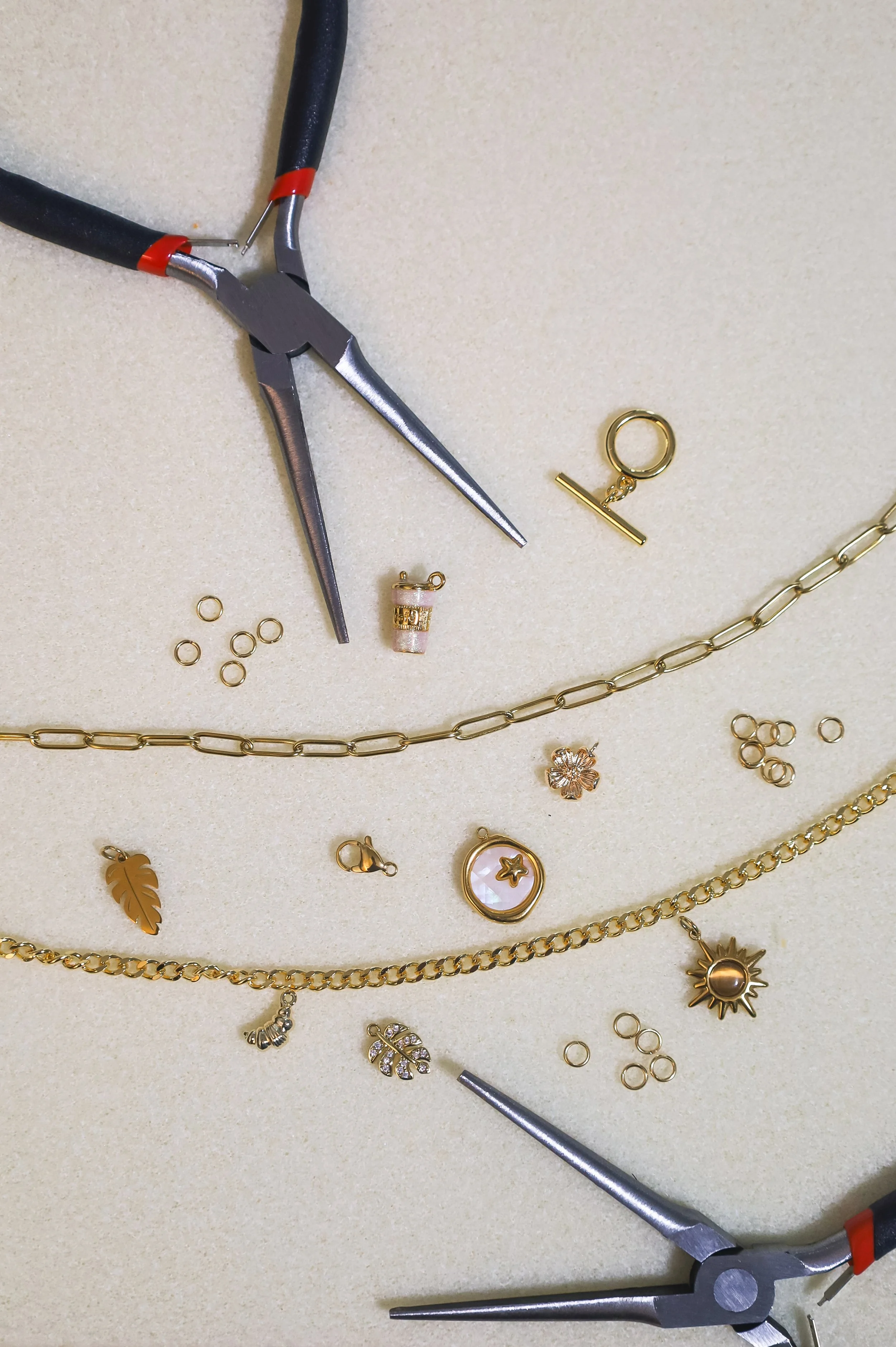 Gold jewelry including necklaces, charms, and rings, along with jewelry pliers on a beige surface.