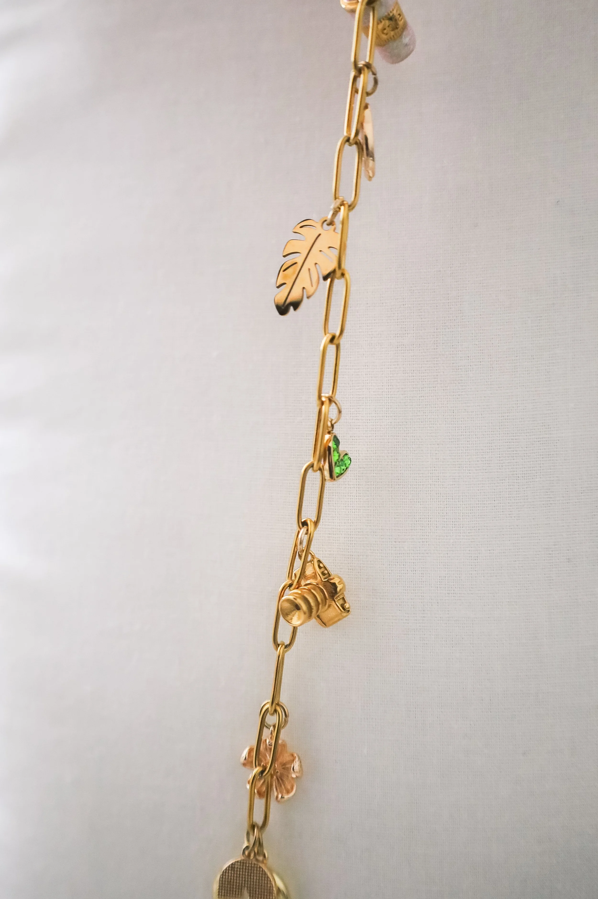 A gold-colored chain necklace with small charms, including a leaf, camera, butterfly, and a green heart, hanging vertically against a plain light background.