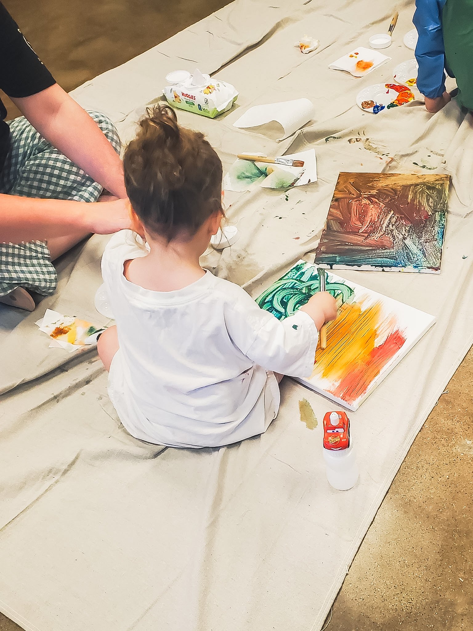 A toddler girl in a white shirt is sitting on the floor painting a colorful abstract artwork on canvas.
