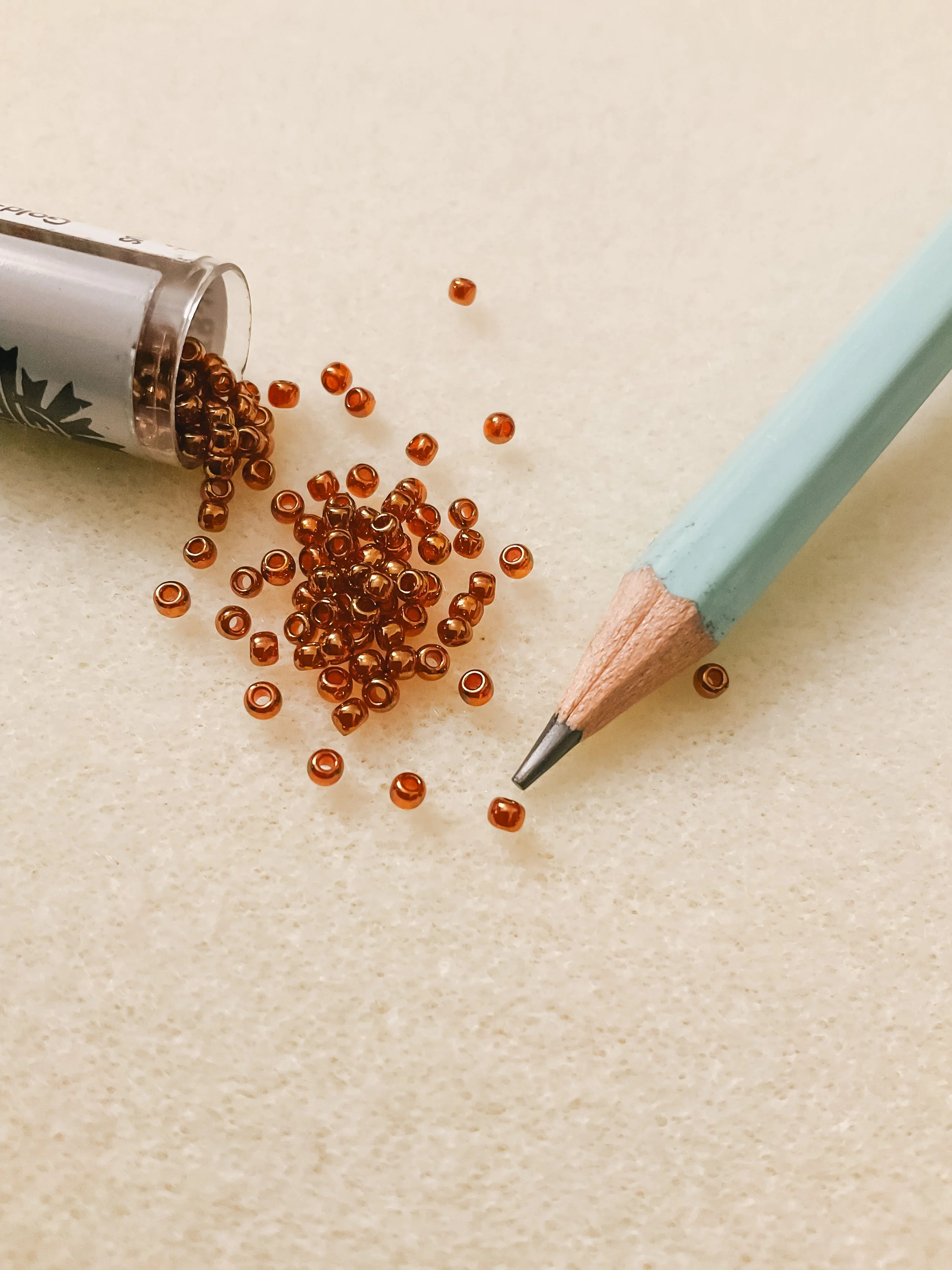 Small brown beads spilled from a clear container onto a light-colored surface, with a light blue pencil nearby.