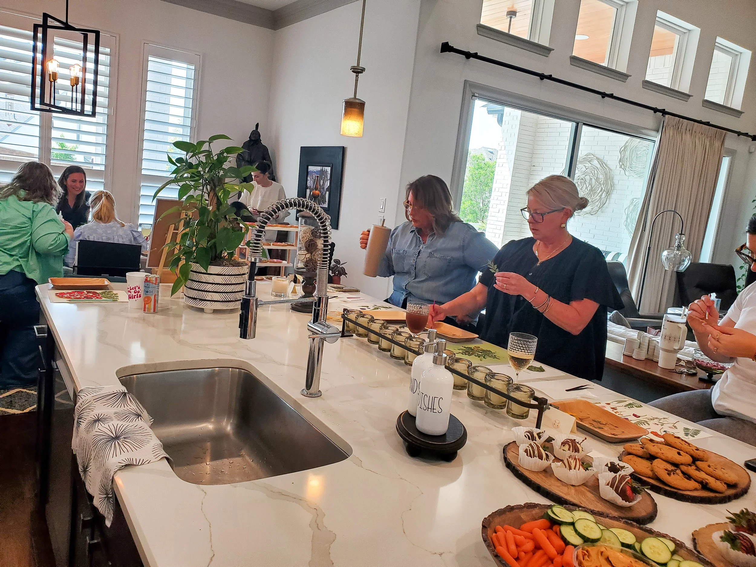 People gathered around a kitchen island with food and drinks, including cookies, vegetables, and beverages, during a pressed flower art party in a well-lit modern kitchen.