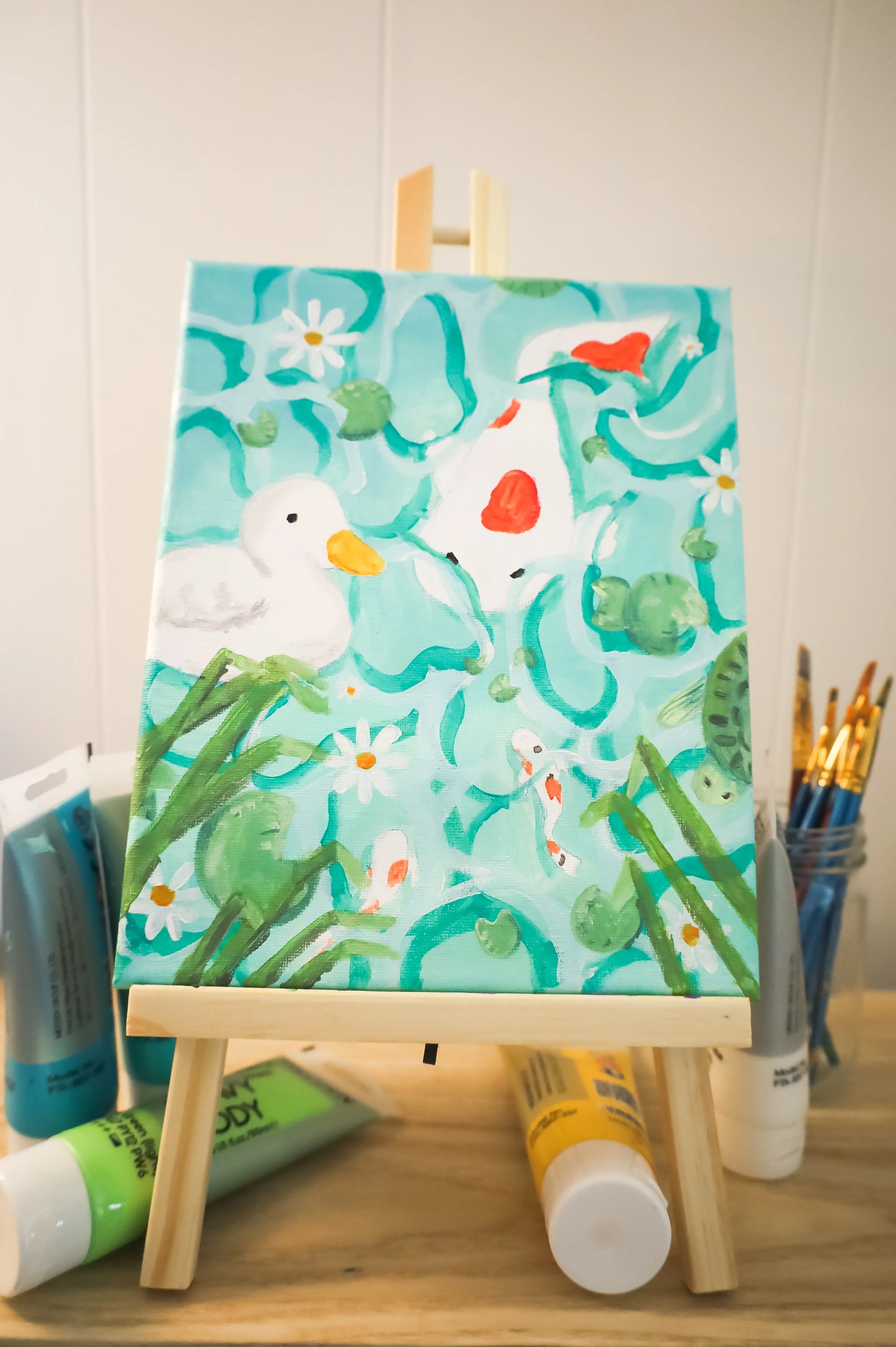 A colorful painting of a pond scene with ducks, fish, lily pads, and flowers on a small easel, surrounded by paint tubes and brushes.