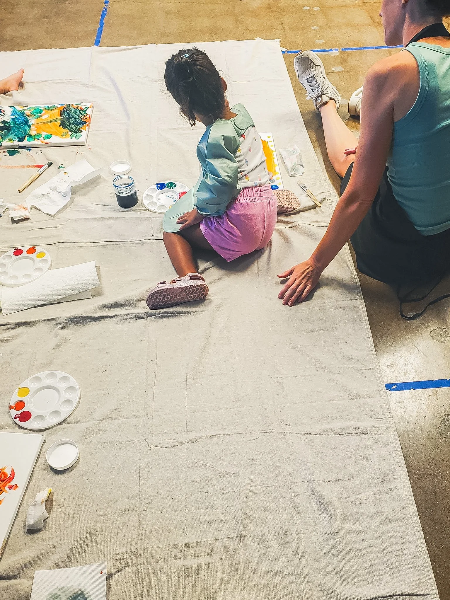 A young girl with dark hair, pink shorts, and a light green smock is sitting on a beige cloth-covered floor painting on canvas. An adult woman, wearing a teal tank top and black shorts, is sitting beside her.