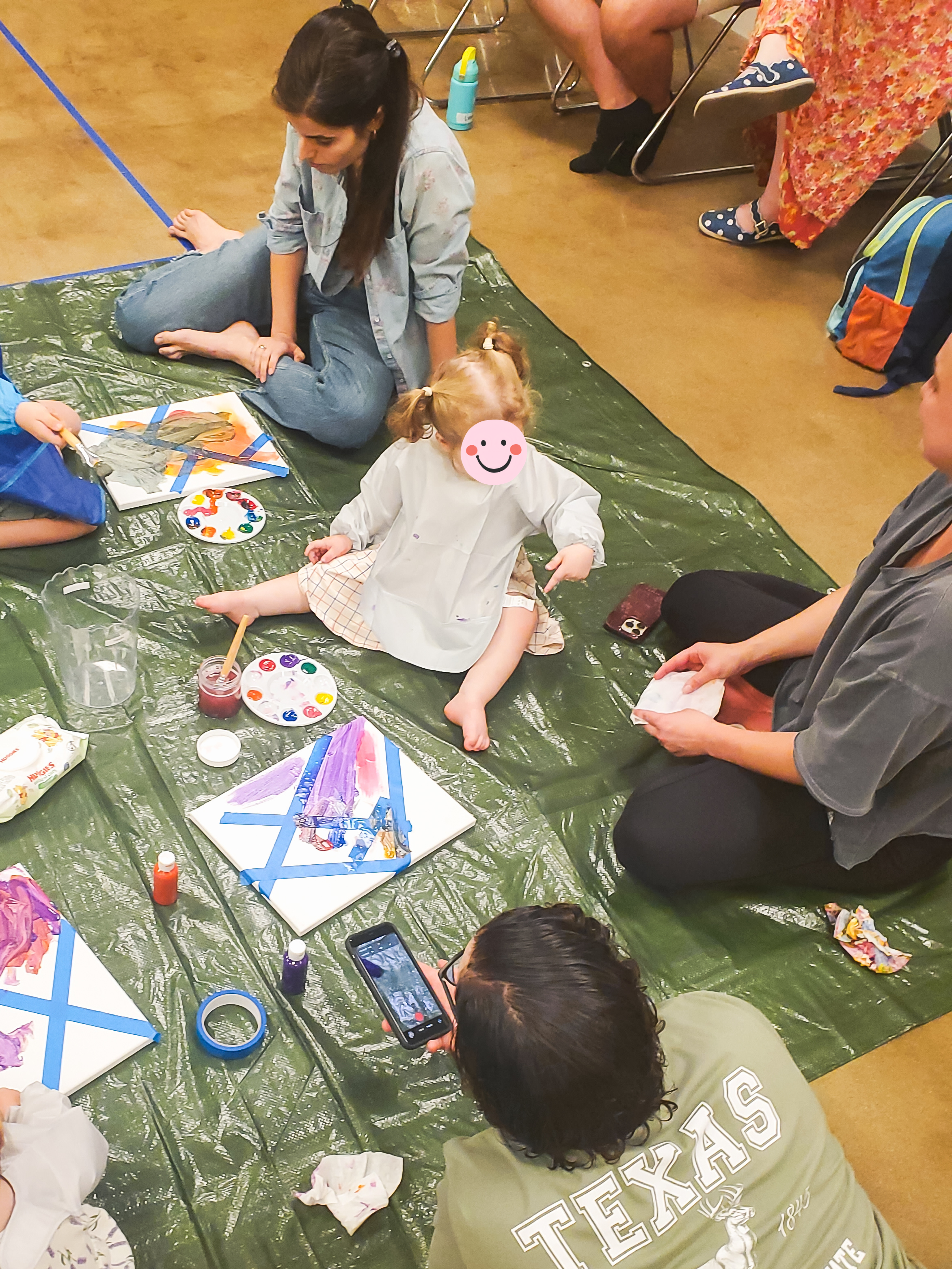 People sitting on the floor, engaged in arts and crafts activities. Art supplies like paint palettes, brushes, and paper are visible on the floor.