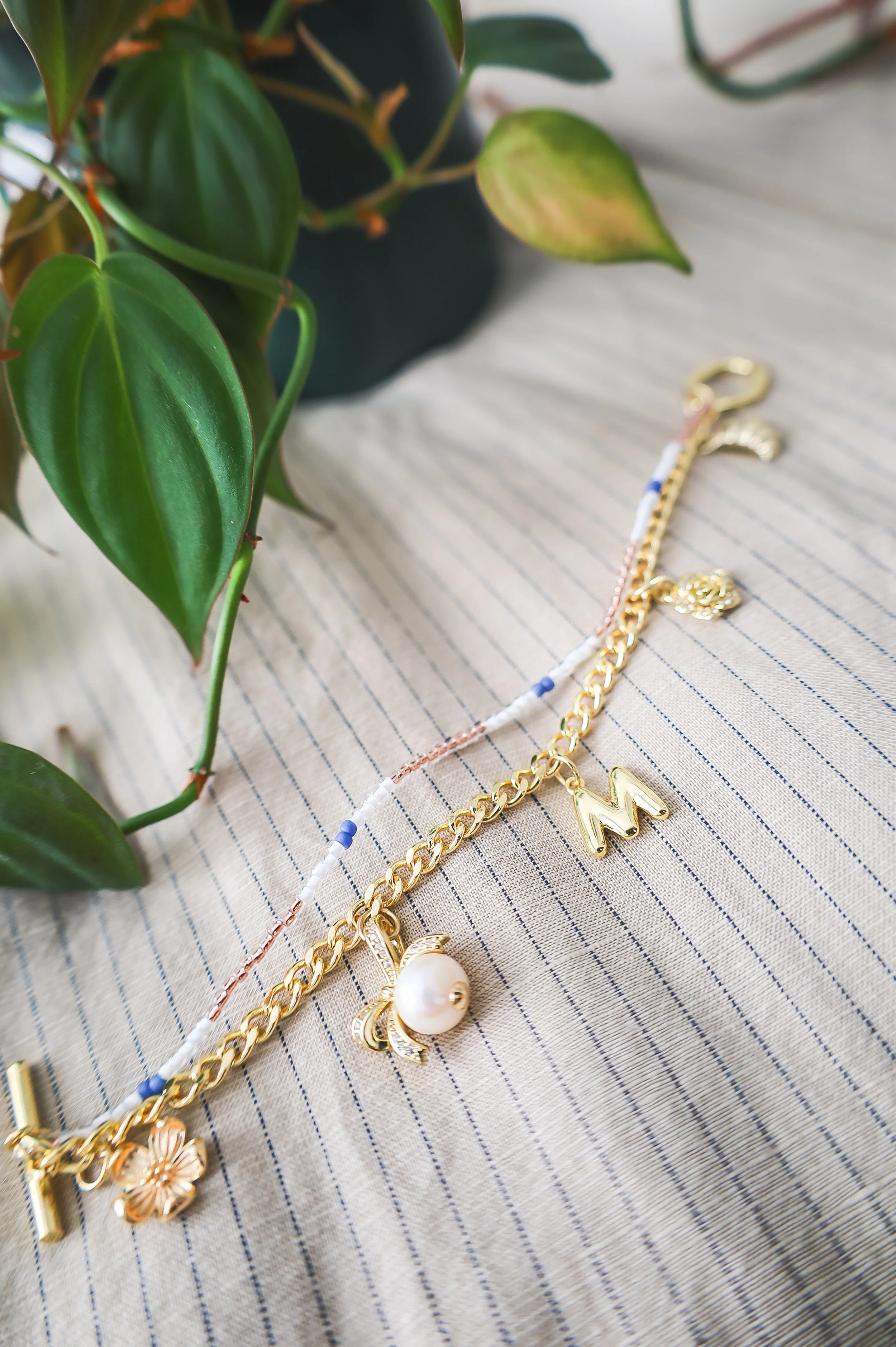Gold chain and charm bracelet on striped fabric surface near green leafy plant.
