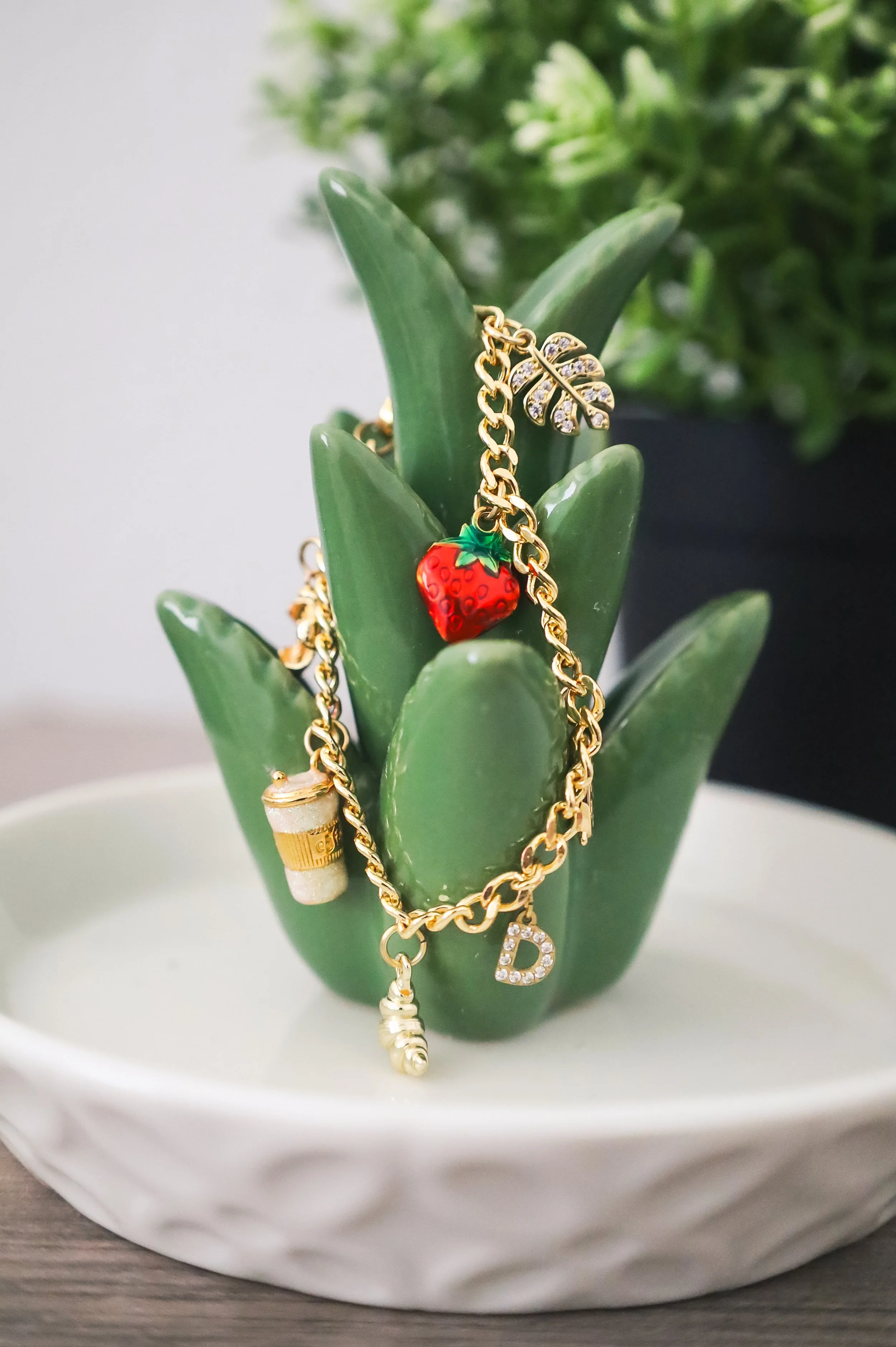 Gold jewelry including a chain, a strawberry charm, a coffee cup charm, a seashell charm, a D charm, and a leaf charm, hanging on a green cactus plant in a white dish.