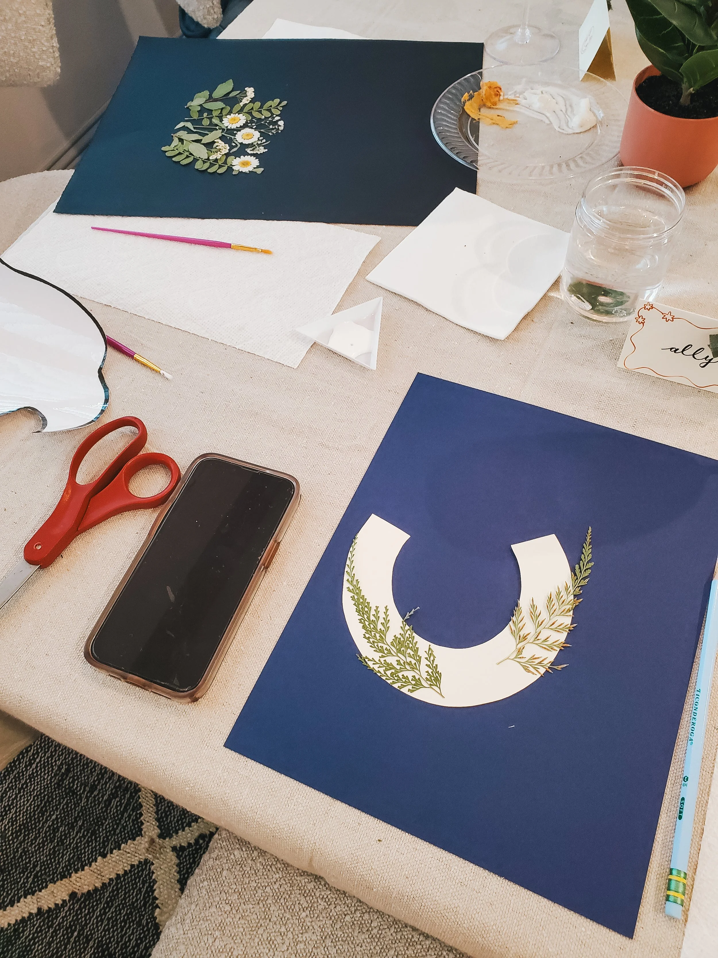 A table with art supplies, including a blue paper with a white C-shape and greenery design, a pair of red scissors, a smartphone, white paper towels, paintbrushes, a glass with water, and decorative items like a plant and food on plates.