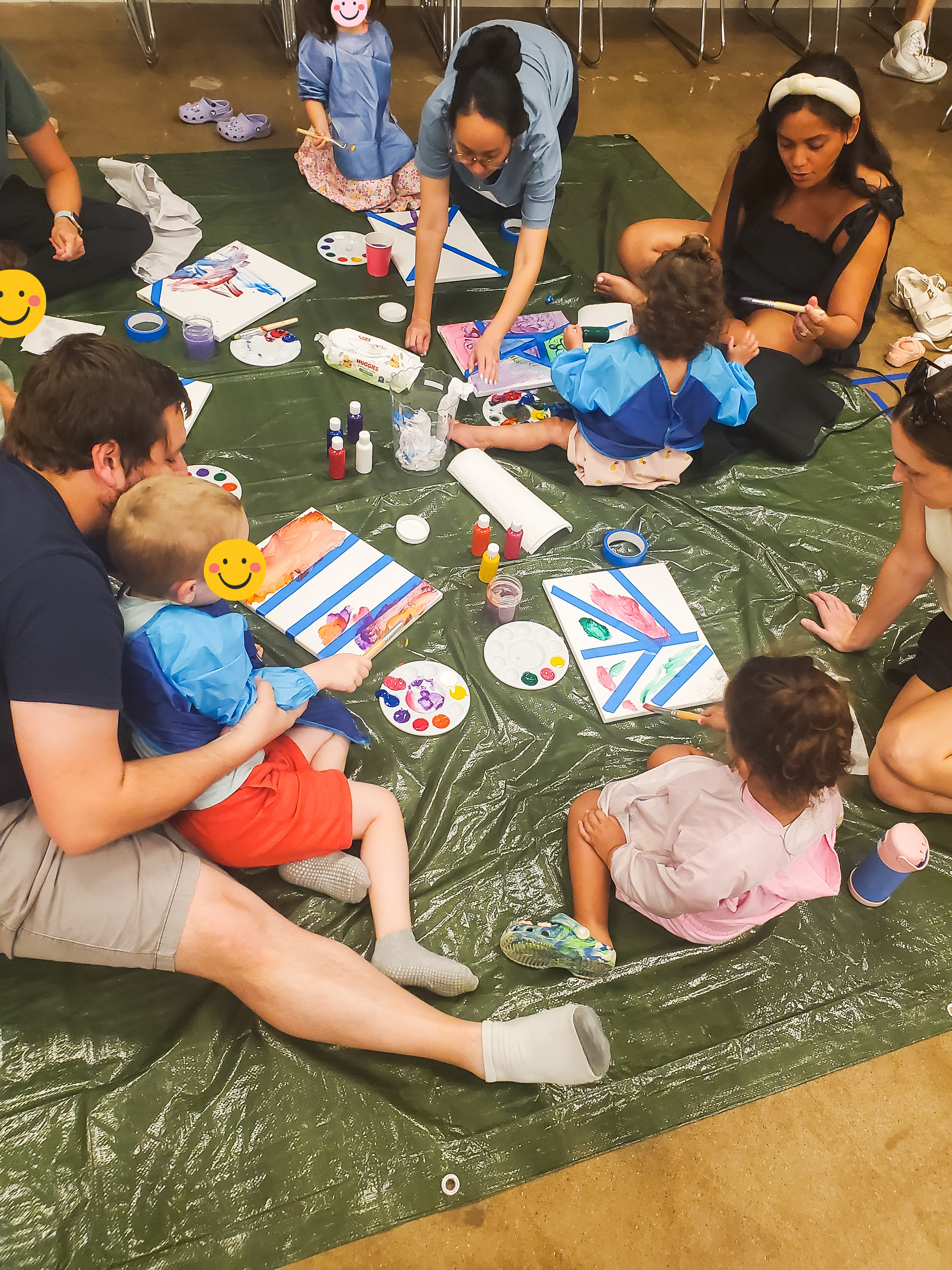 A group of children and adults are sitting on a green tarp-covered floor, painting on canvases with watercolors. The children are wearing smocks, and there are various painting supplies scattered around.