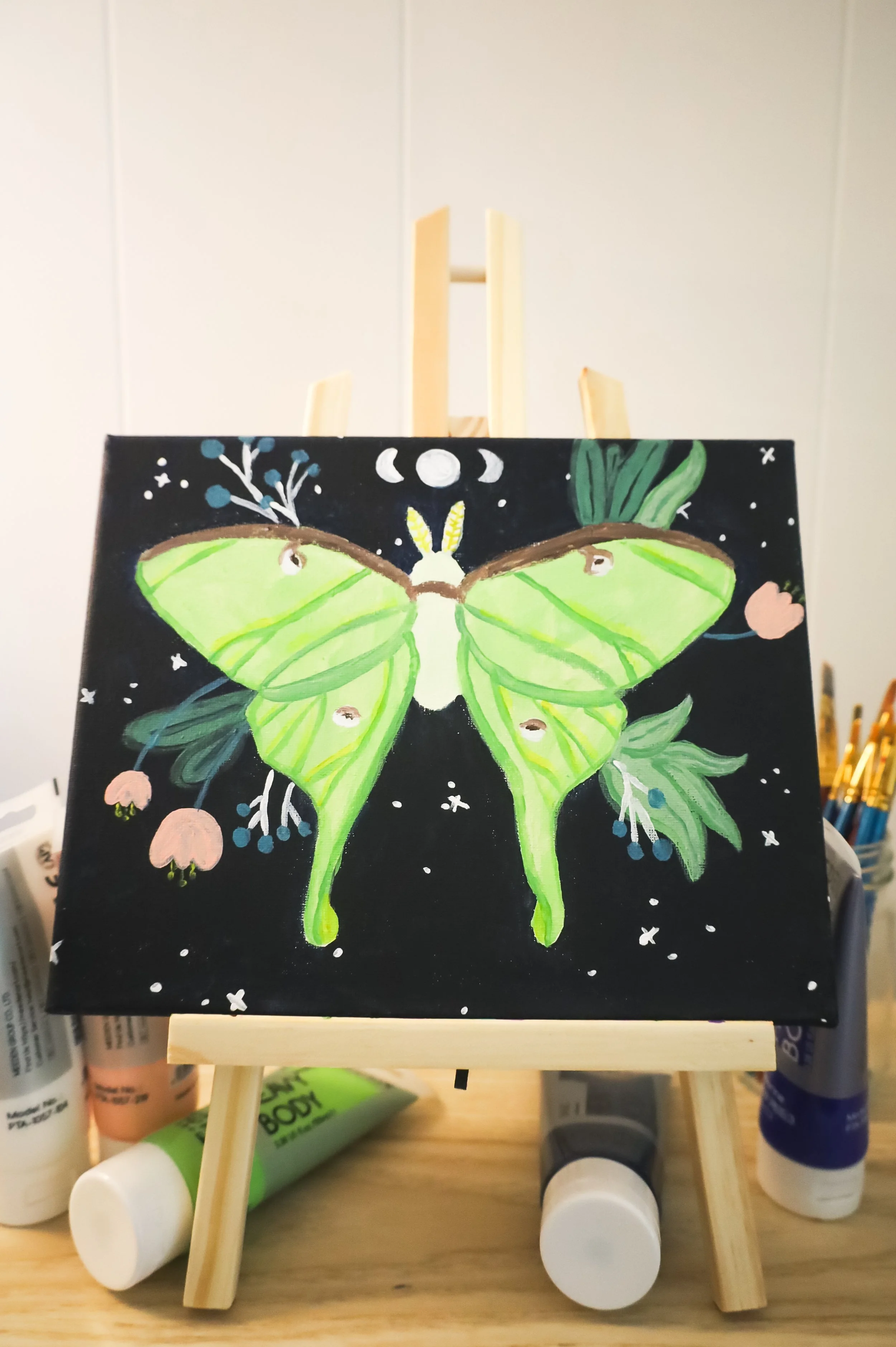 A painting of a green moth with floral and moon designs on a black background, displayed on a small wooden easel.