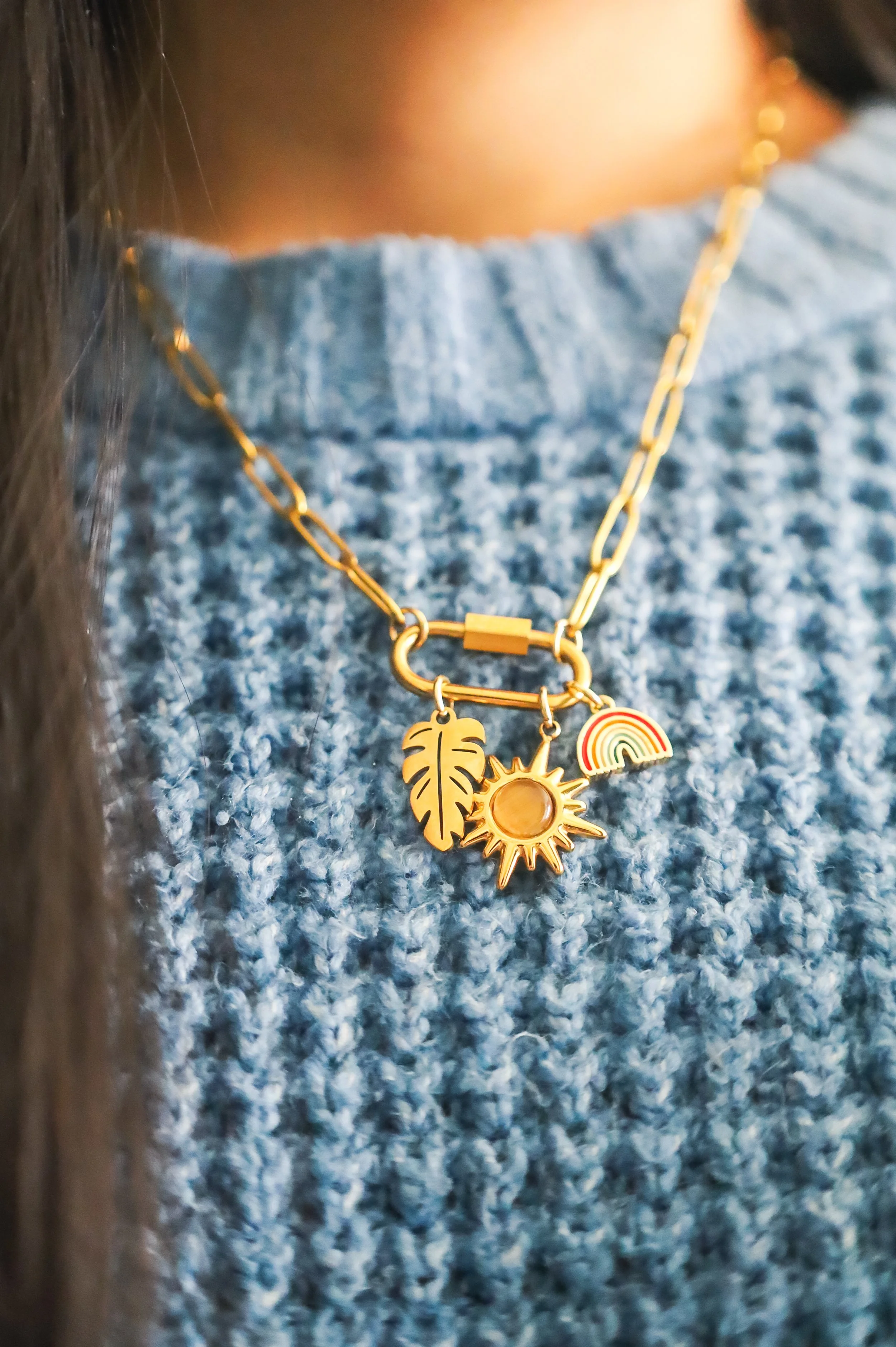 Close-up of a gold necklace with three charms: a leaf, a sun, and a rainbow, worn over a textured blue sweater.