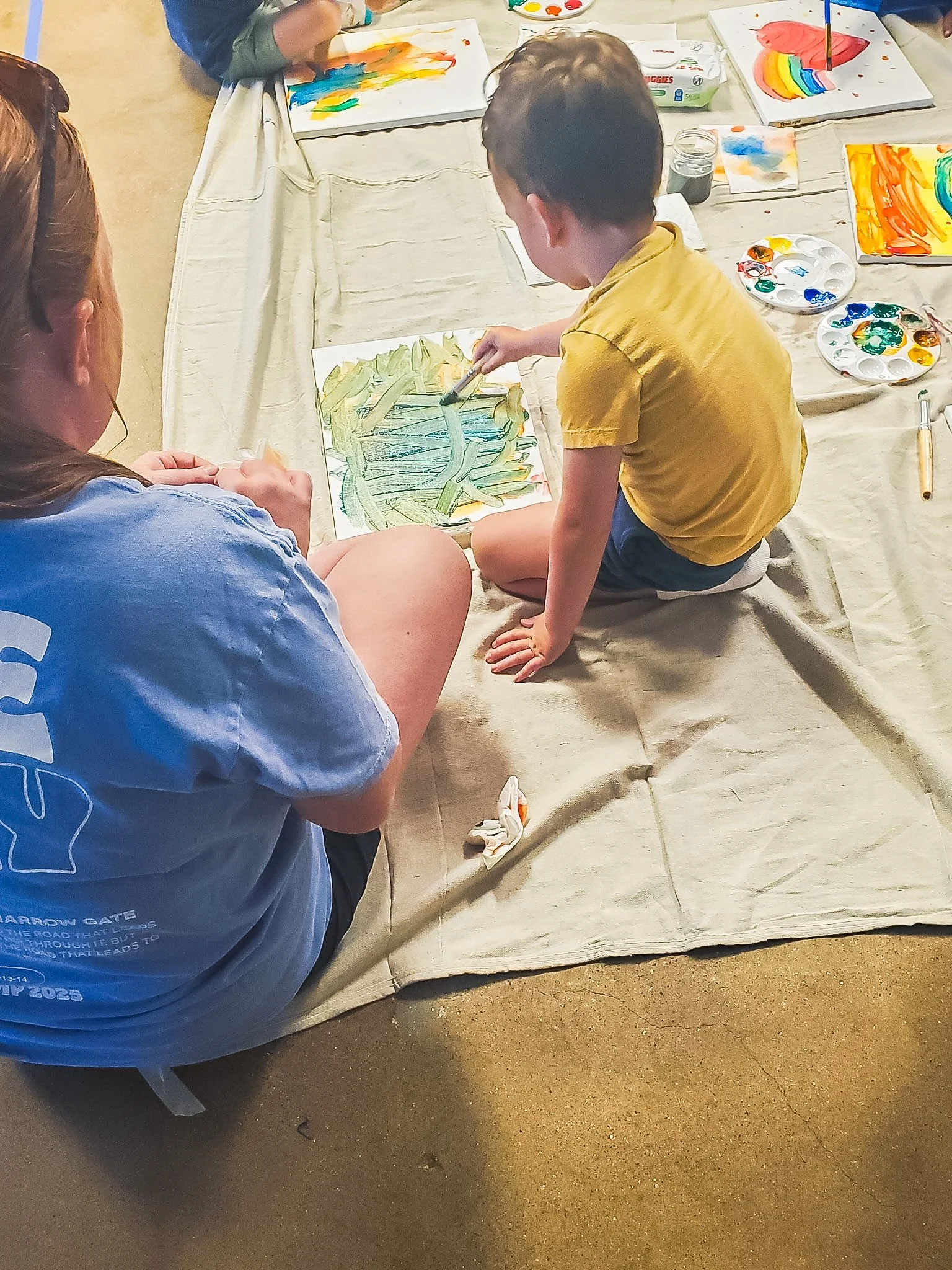 Child and an adult sitting on the floor, painting on a canvas. The child is focused on their artwork, using a brushes and various colors.