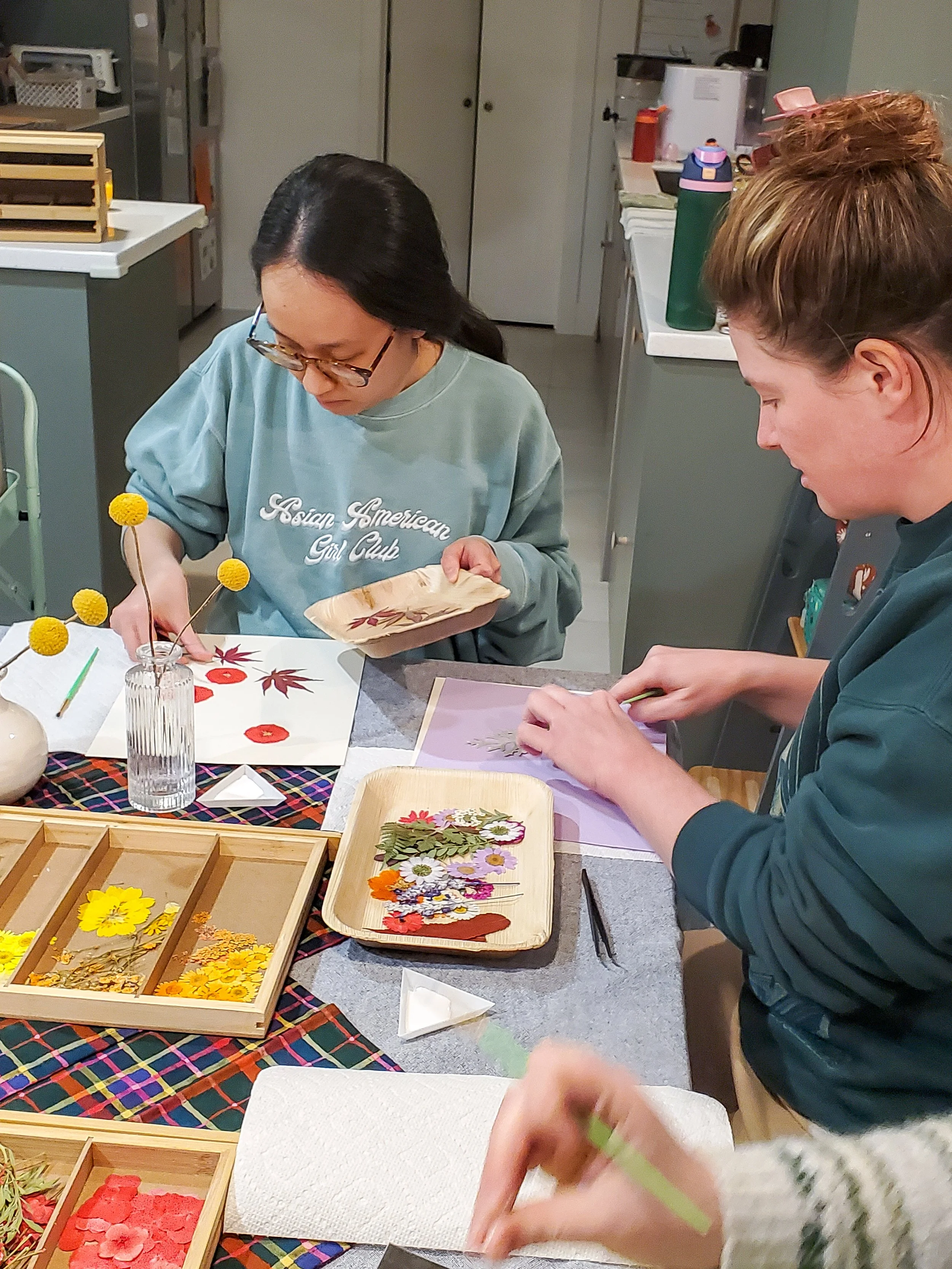 Two women working on craft projects at a table. One woman, wearing glasses and a sweatshirt that says 'Asian American Girl Club,' is placing yellow pom-poms on a paper. The other woman is arranging flowers or leaves on a paper. The table has supplies like flowers, leaves, and craft materials, with a tray of yellow flowers and a glass of water nearby.